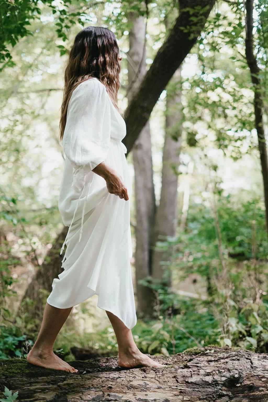 A woman in a flowing white dress walking barefoot on a fallen tree trunk in a lush green forest. Experiencing healing through somatic coaching.
