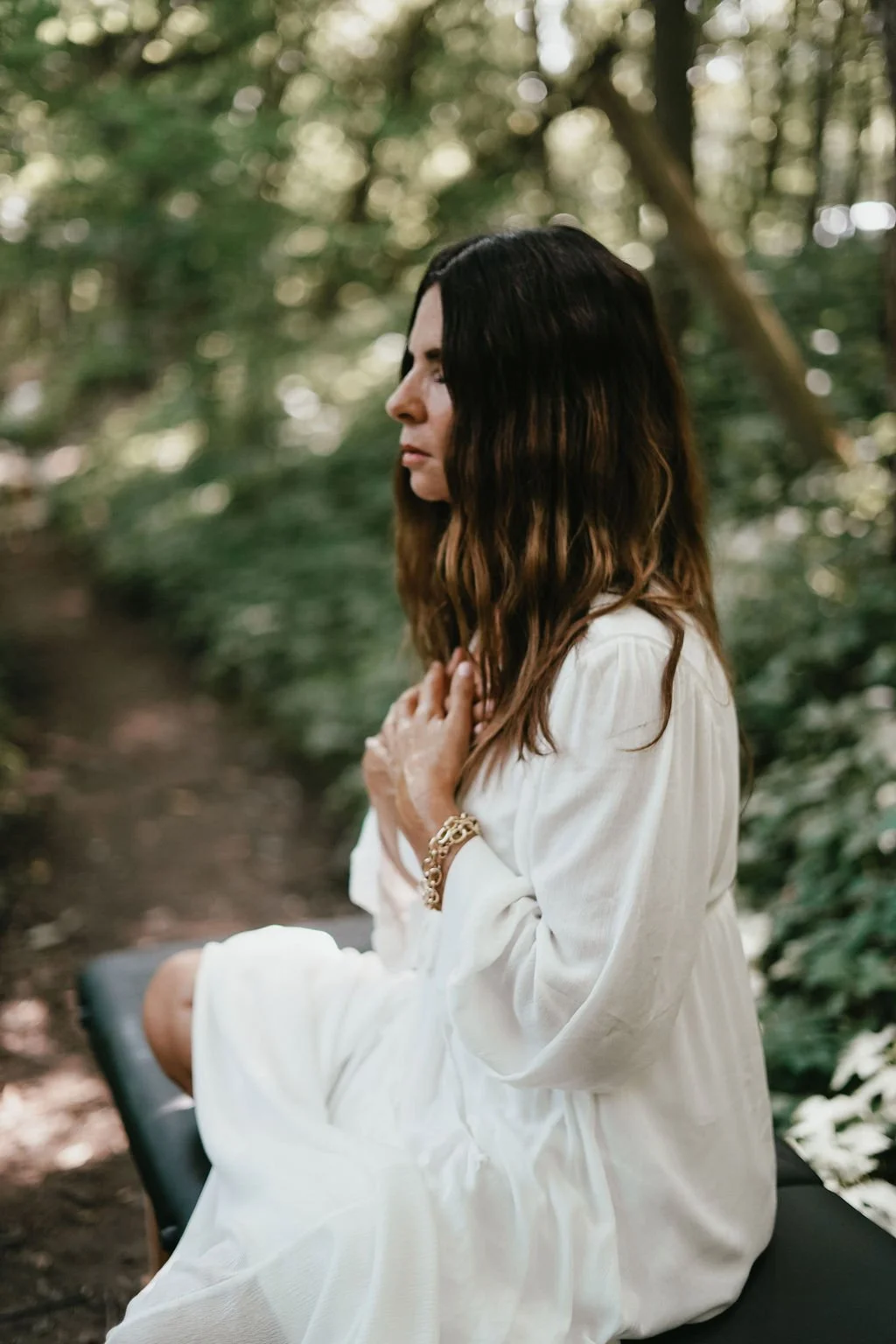 A woman sitting outdoors in a wooded area, wearing a white dress and jewelry, with her eyes closed and hands on her chest. Experiencing healing through somatic coaching.