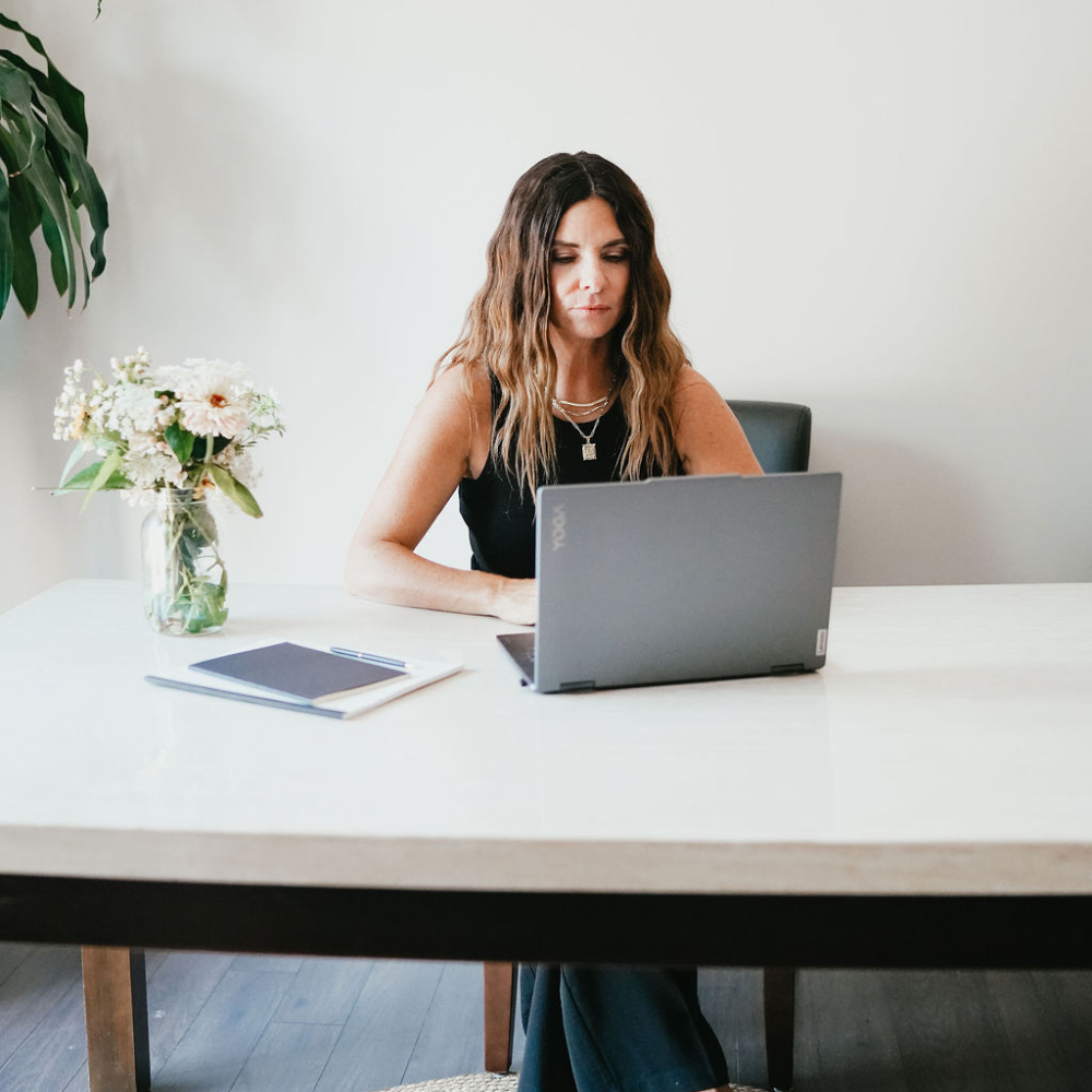 A woman with wavy brown hair sitting at a white desk, working on a gray laptop. There is a vase of white and pink flowers, a blue notebook, and a pen on the desk. The background is plain with a large green plant fixed on the wall to the left.