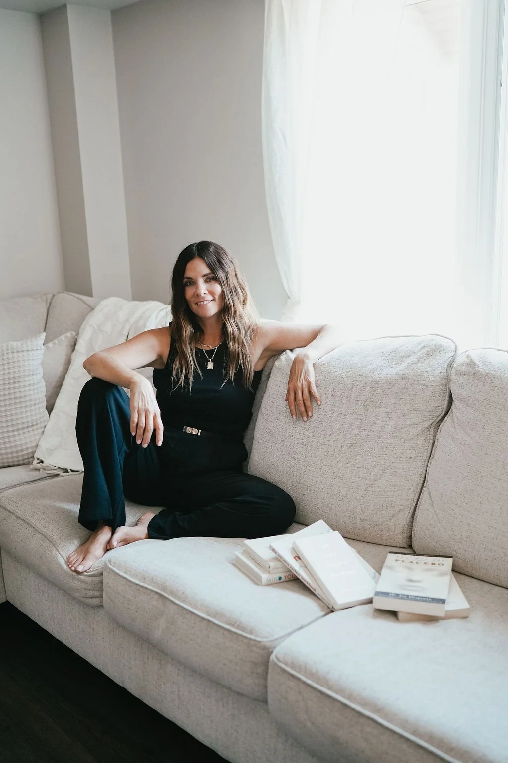Woman sitting on a light-colored sofa next to a window, wearing a black outfit, with books placed on the sofa in front of her.
