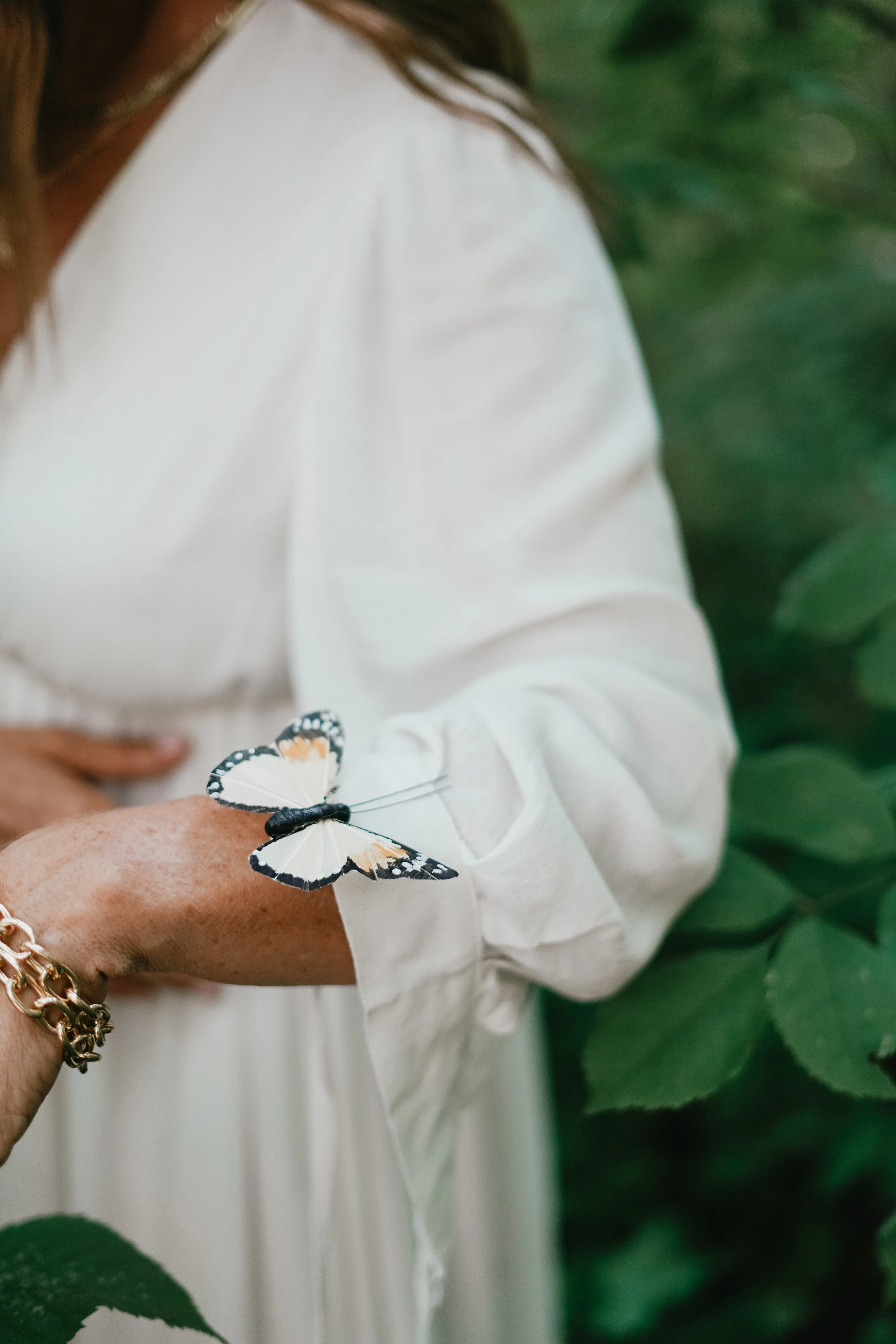 Person wearing a white dress with a butterfly resting on their arm, surrounded by green foliage. Experiencing healing through somatic coaching.