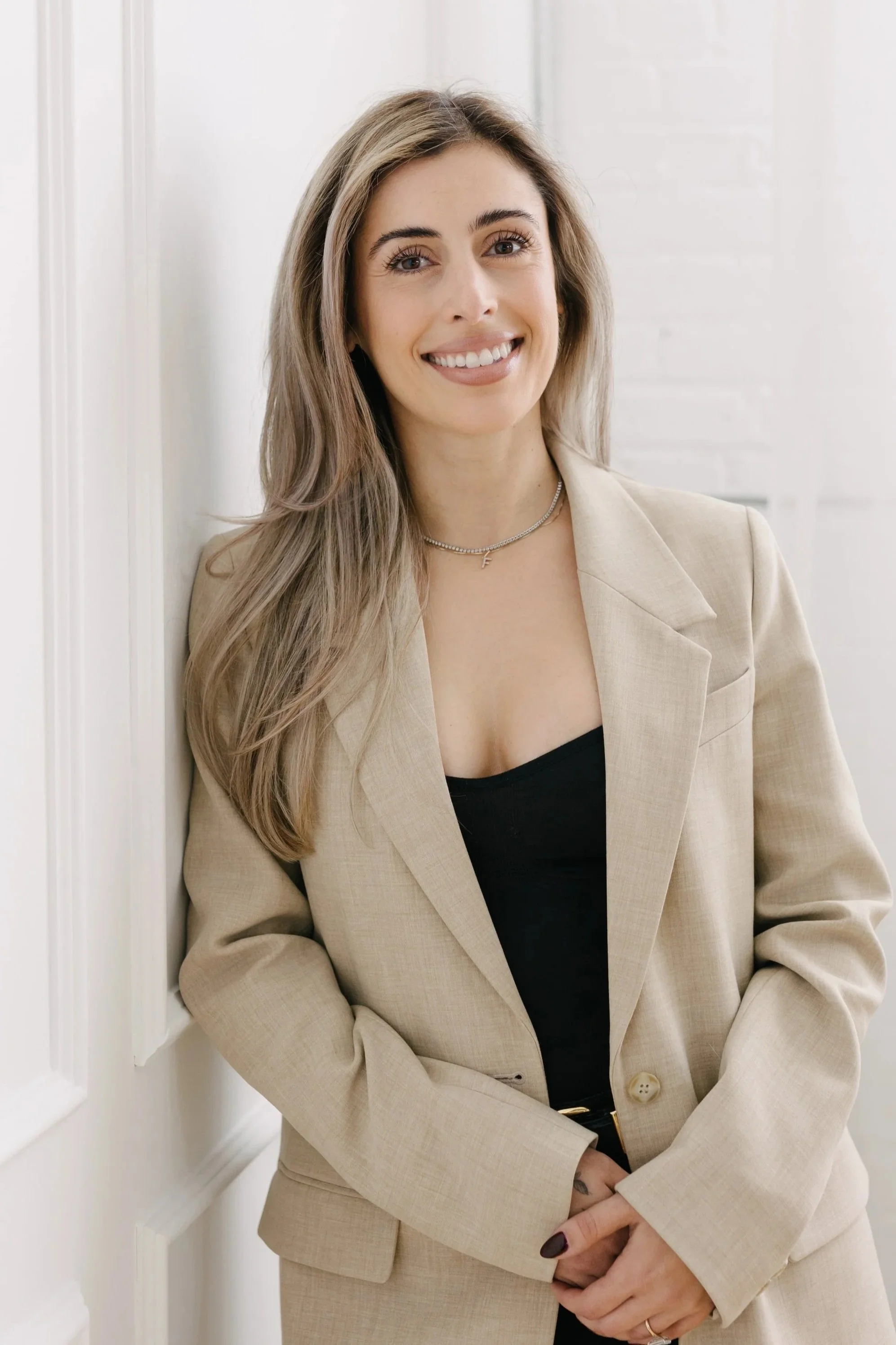 A woman with long, light brown hair and light skin leaning against a wall, smiling at the camera, wearing a beige blazer over a black top, with a necklace and dark nail polish.