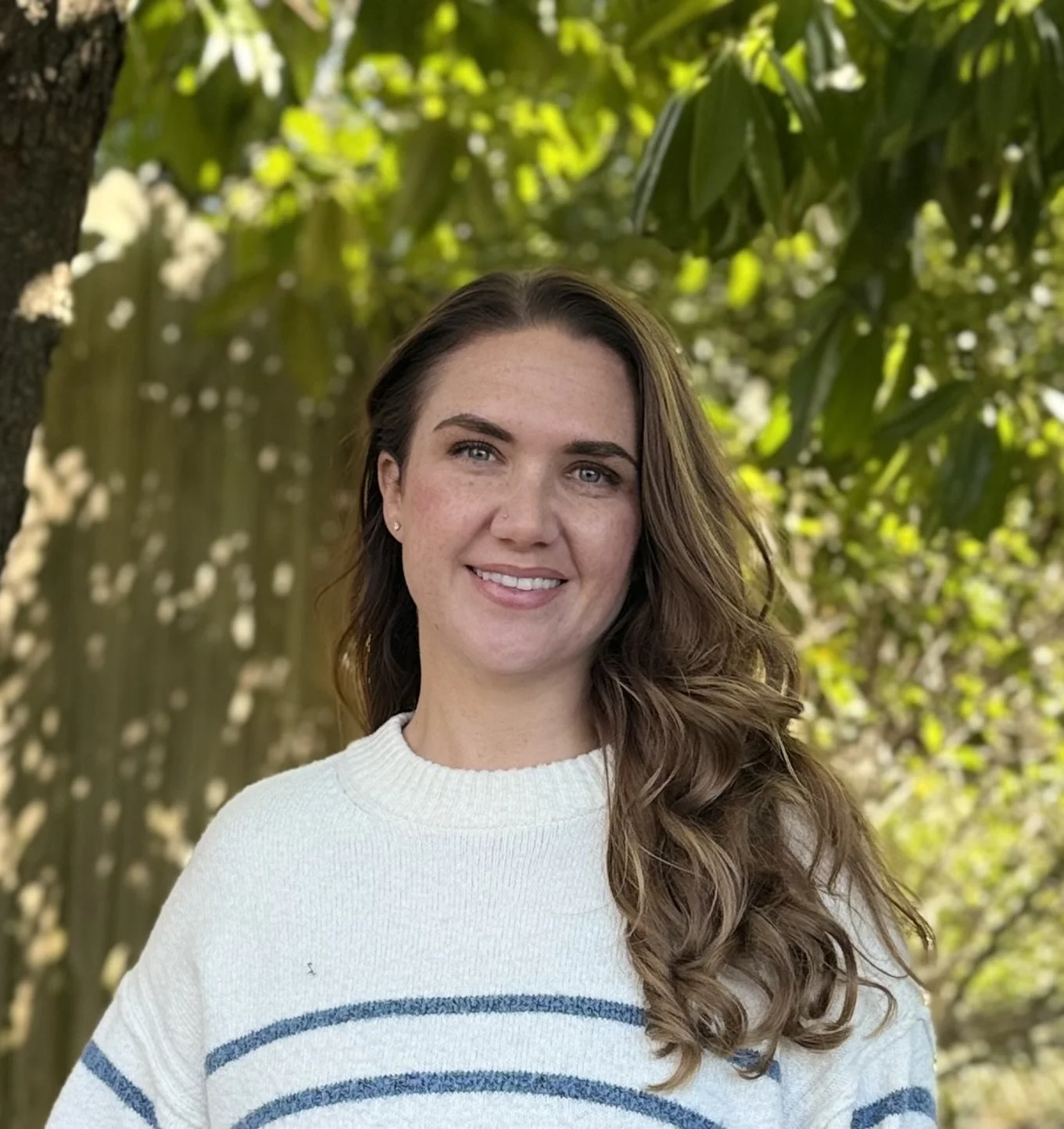A young woman with long, wavy brown hair, blue eyes, and light skin, smiling outdoors with a leafy green background.