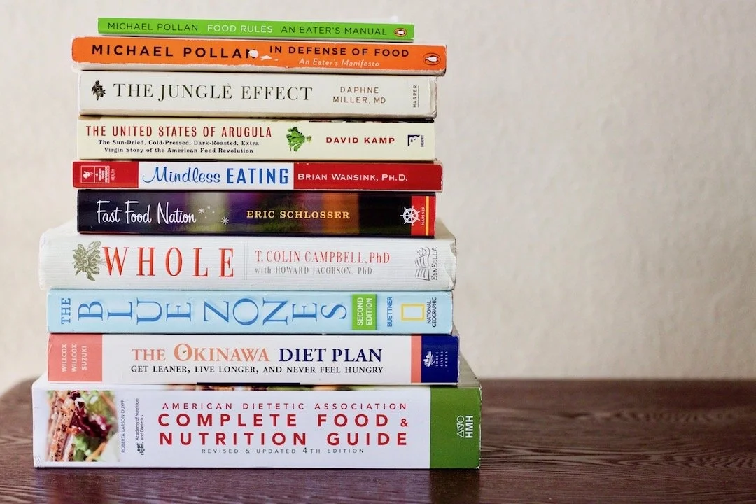Stack of twelve books related to food, nutrition, and eating habits on a wooden table, against a white wall.