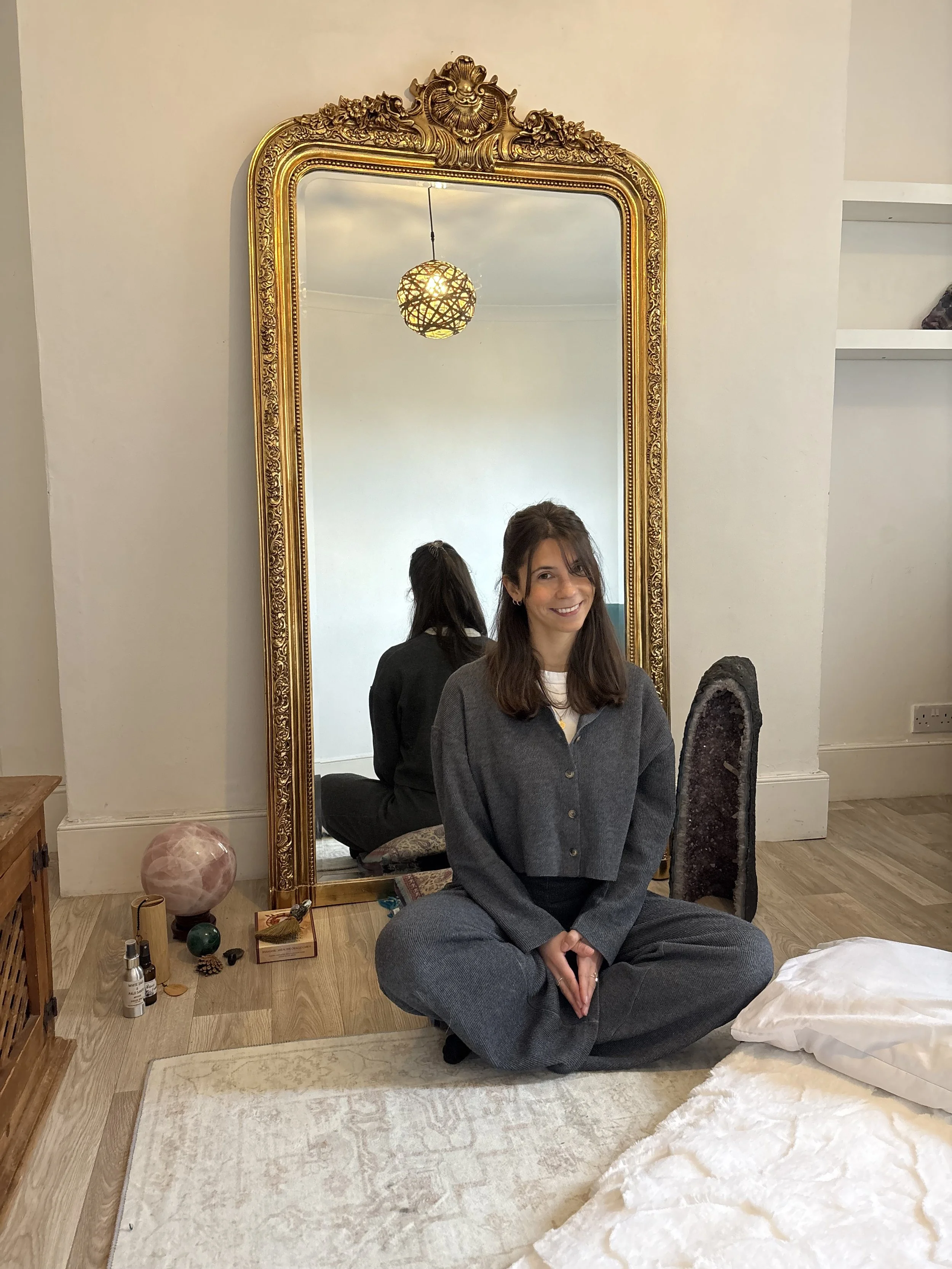 A woman is sitting cross-legged on a beige rug in front of a large, ornate gold-framed mirror. She is smiling, wearing gray baggy pants and a gray jacket. In the reflection, the back of her head is visible. The room has a wooden floor, a small side table with various objects, and a geometric pendant light hanging from the ceiling.