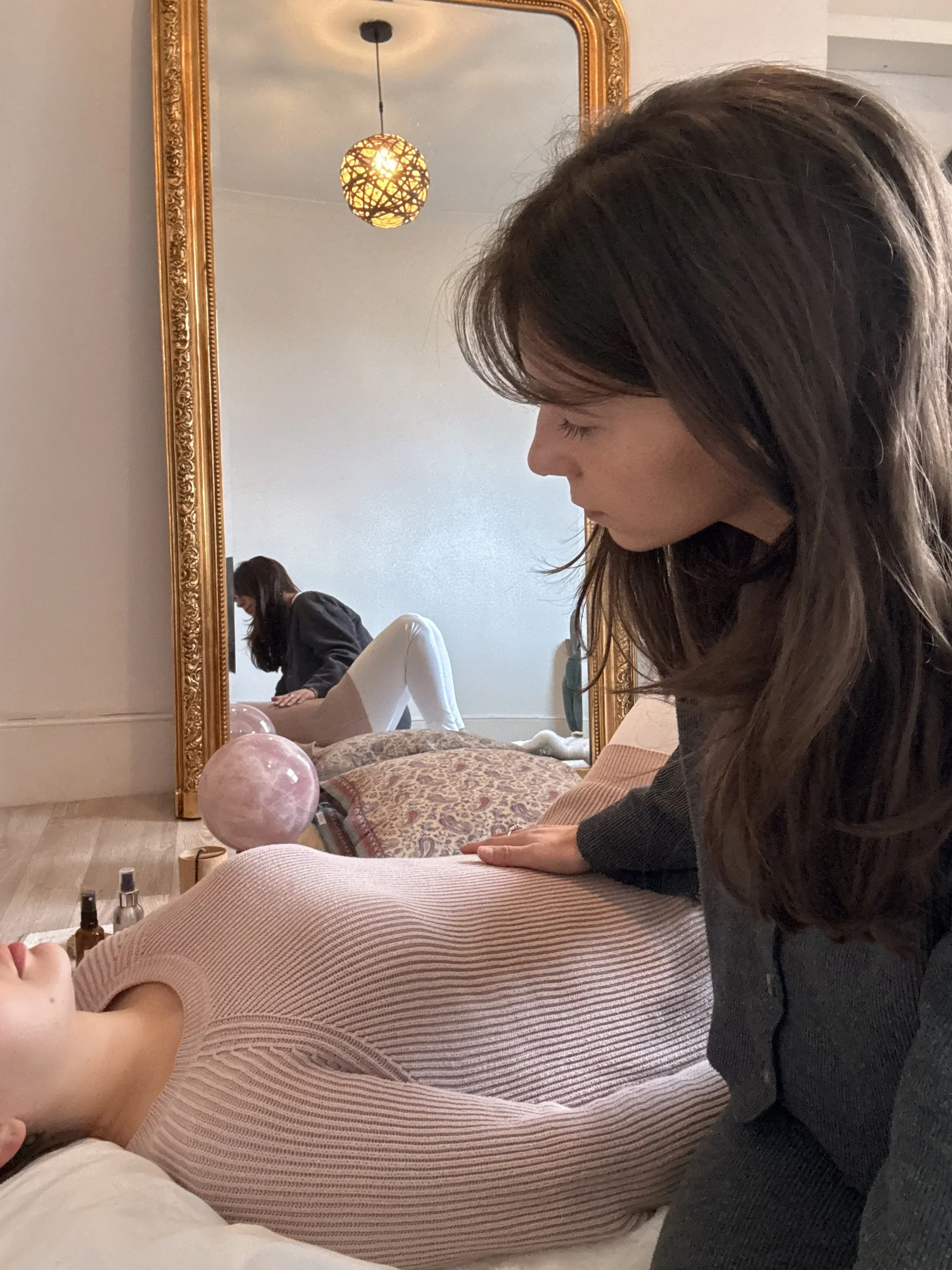 A woman receiving a massage from a massage therapist in a cozy room with a large gold-framed mirror, decorative pillows, pink Himalayan salt lamp, and massage oils.