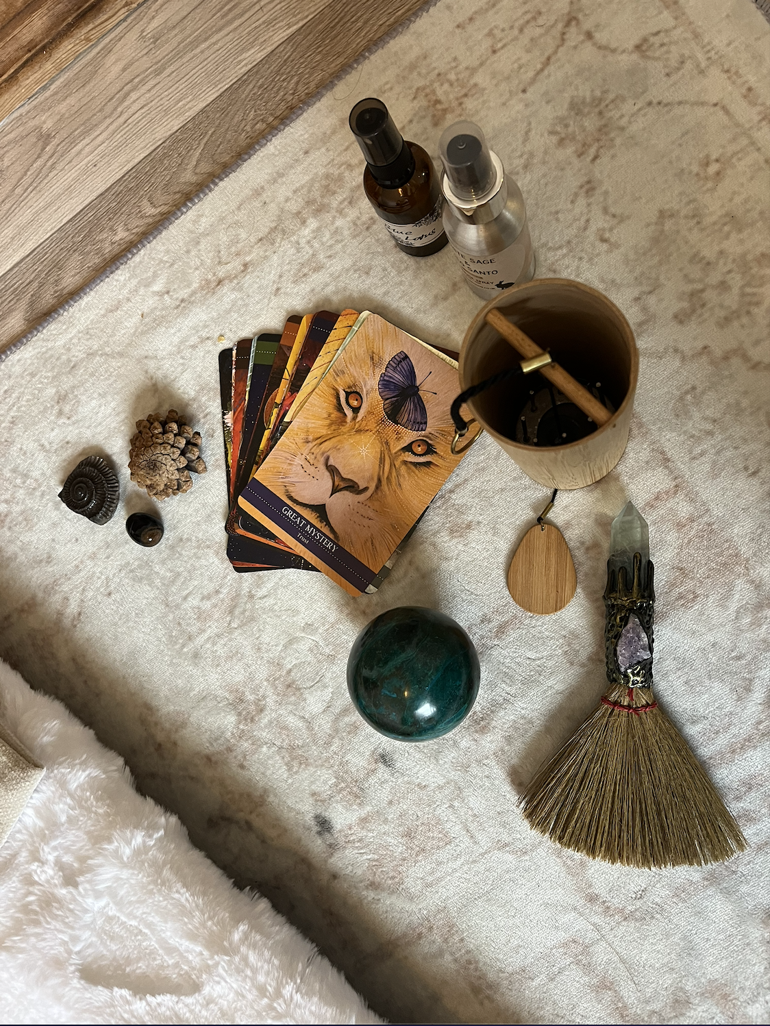 A collection of tarot or oracle cards, crystals, a wooden brush, a sphere, bottles of spray, and a container with a stick inside, arranged on a beige surface.