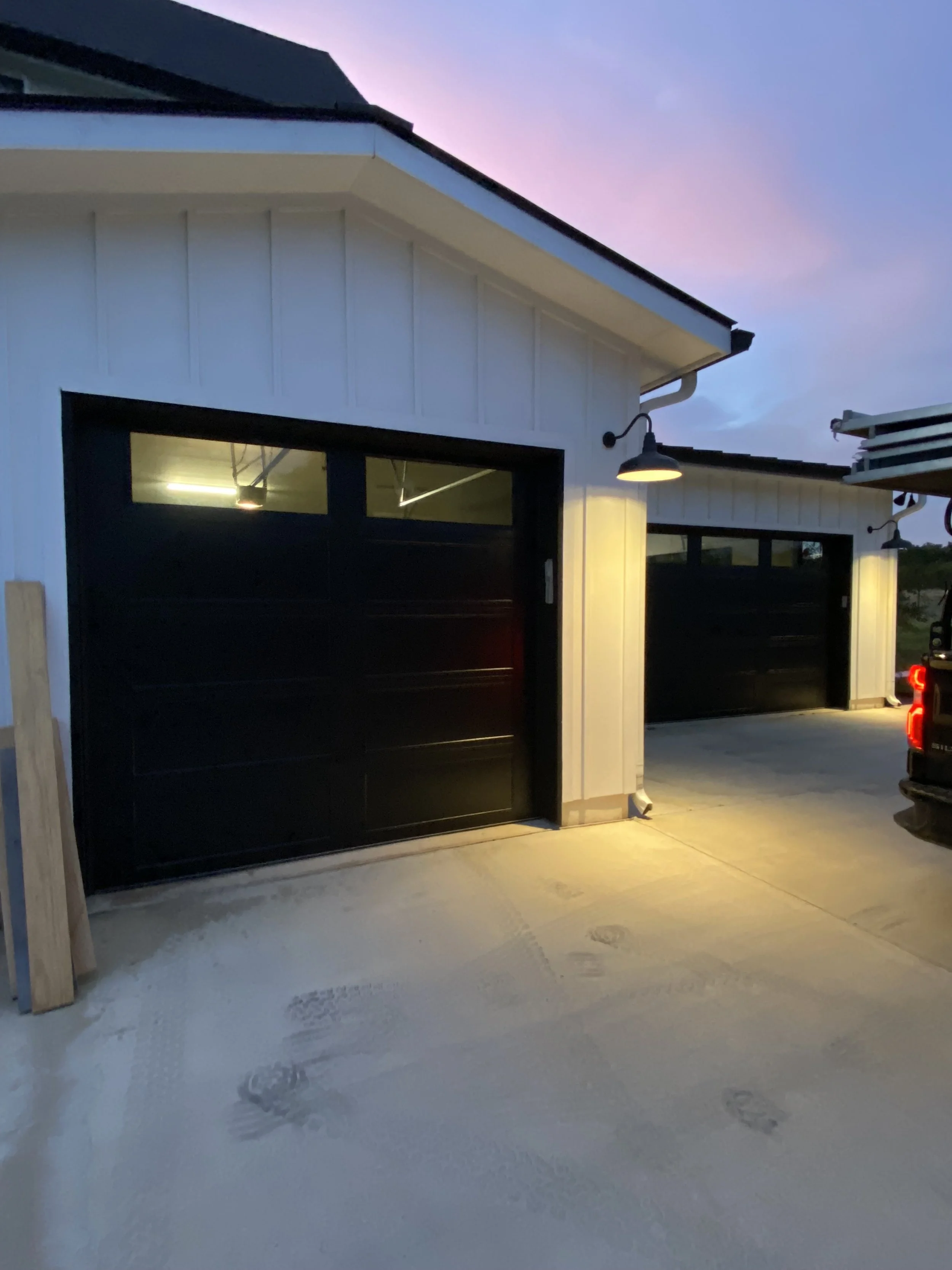 Two 8 foot tall black garage doors with windows on a white modern house with outdoor lights turned on at dusk.