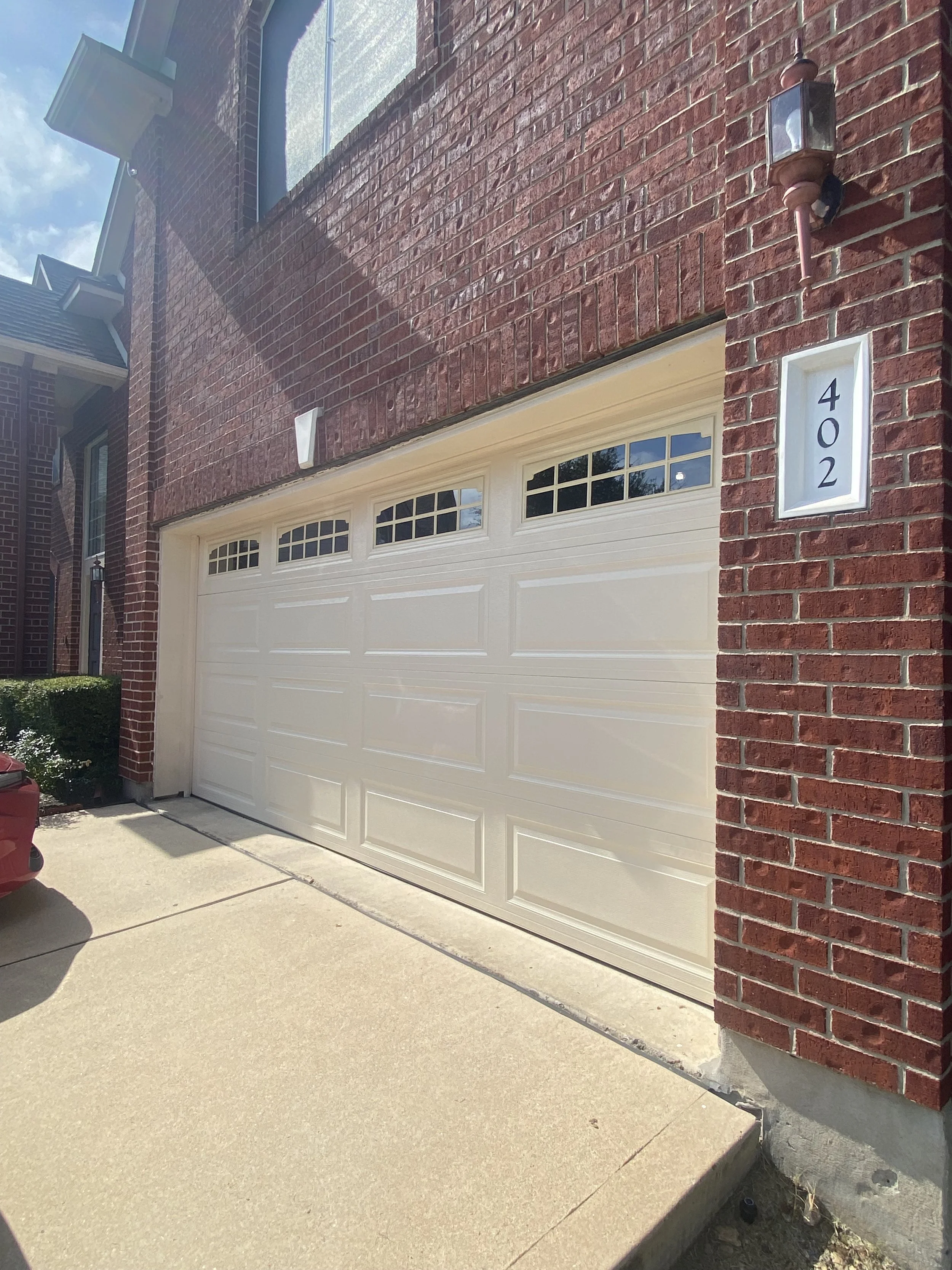 A tan insulated garage door with 4 windows spanning across the top of the door. The windows have inserts giving them a unique design and the door is installed on a brick house.