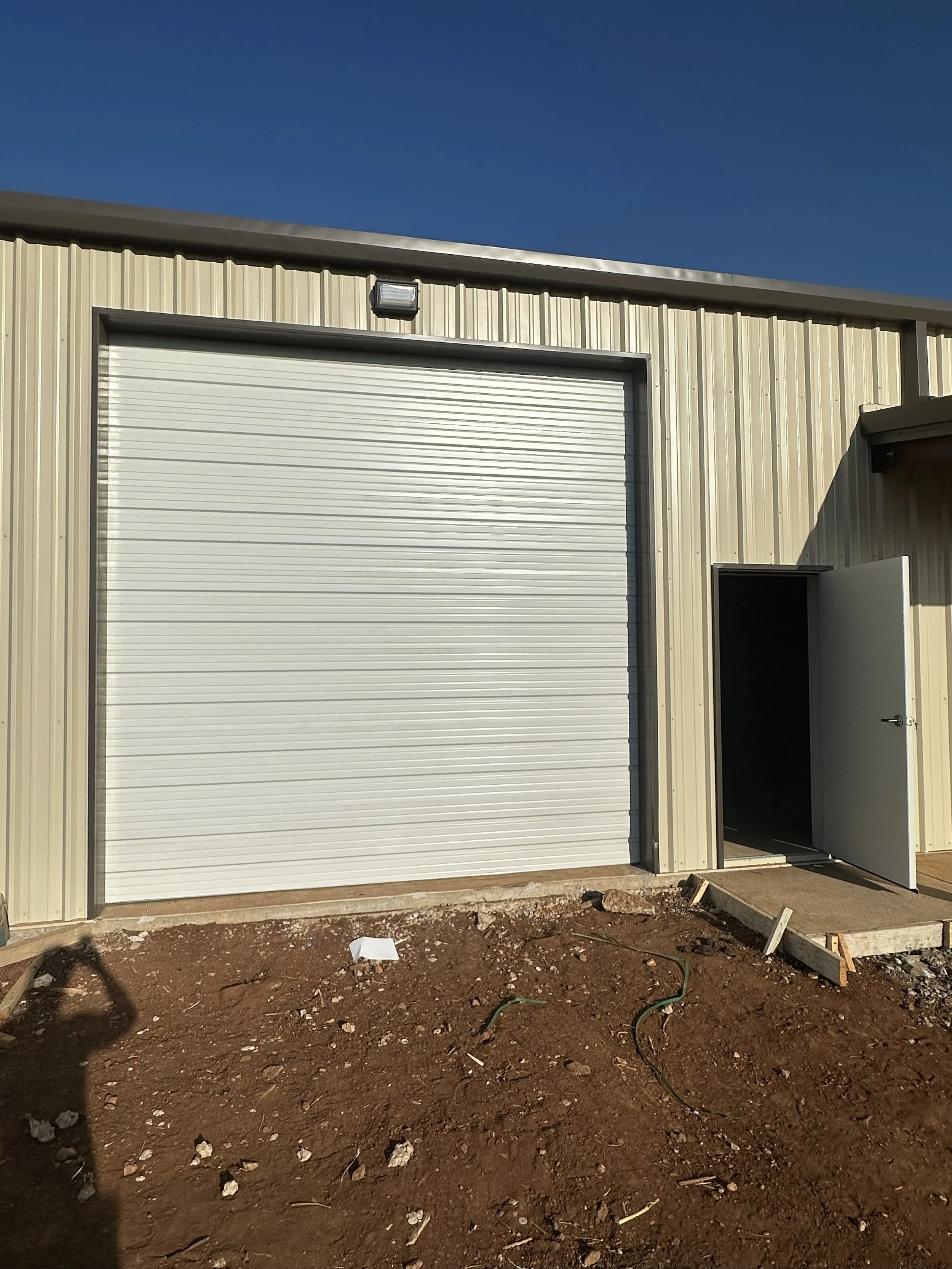 Exterior of a beige metal building with a large closed white roll-up garage door and a smaller open personnel white door to the right, on a dirt ground with construction debris, under a blue sky.