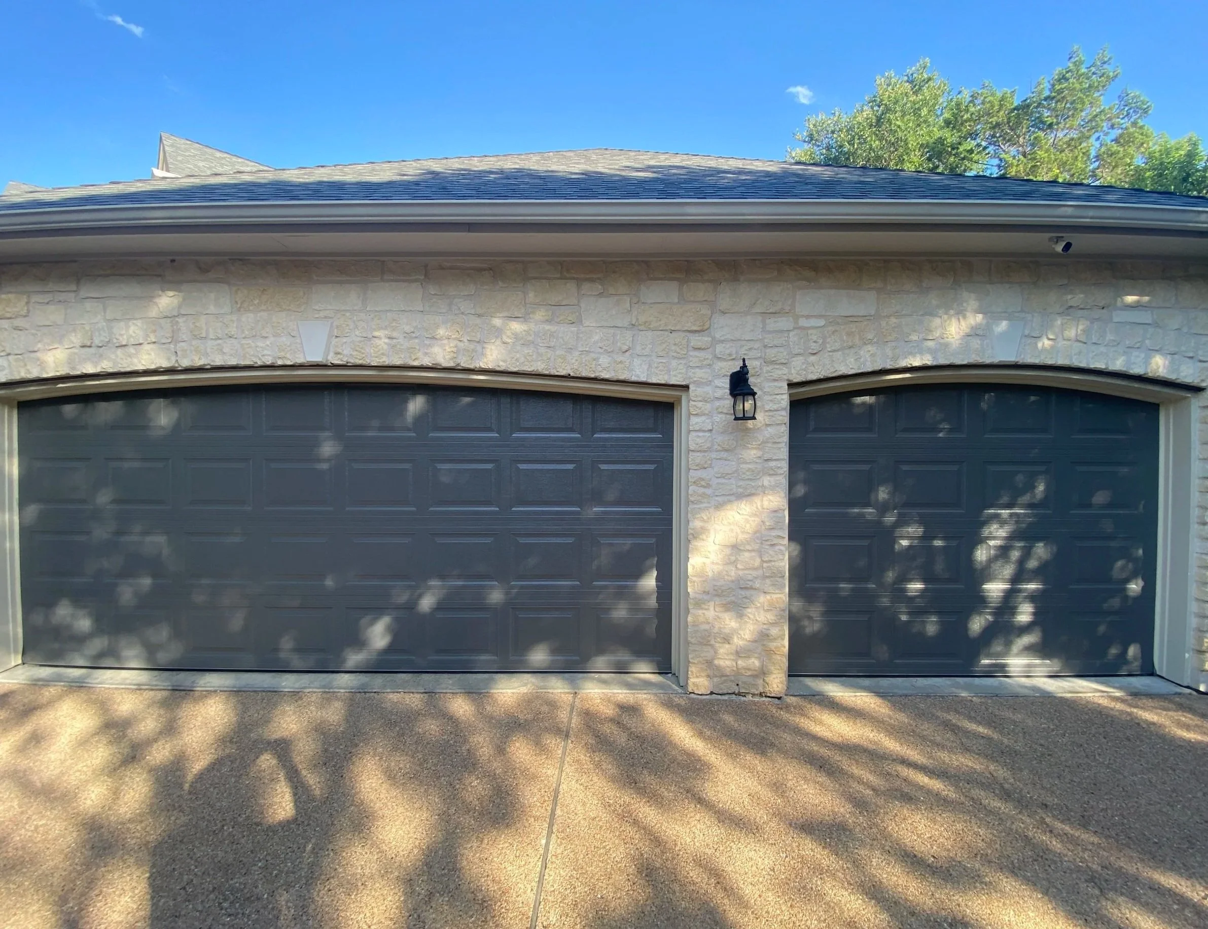 Double charcoal gray garage doors on a stone house with exterior wall lantern and driveway.