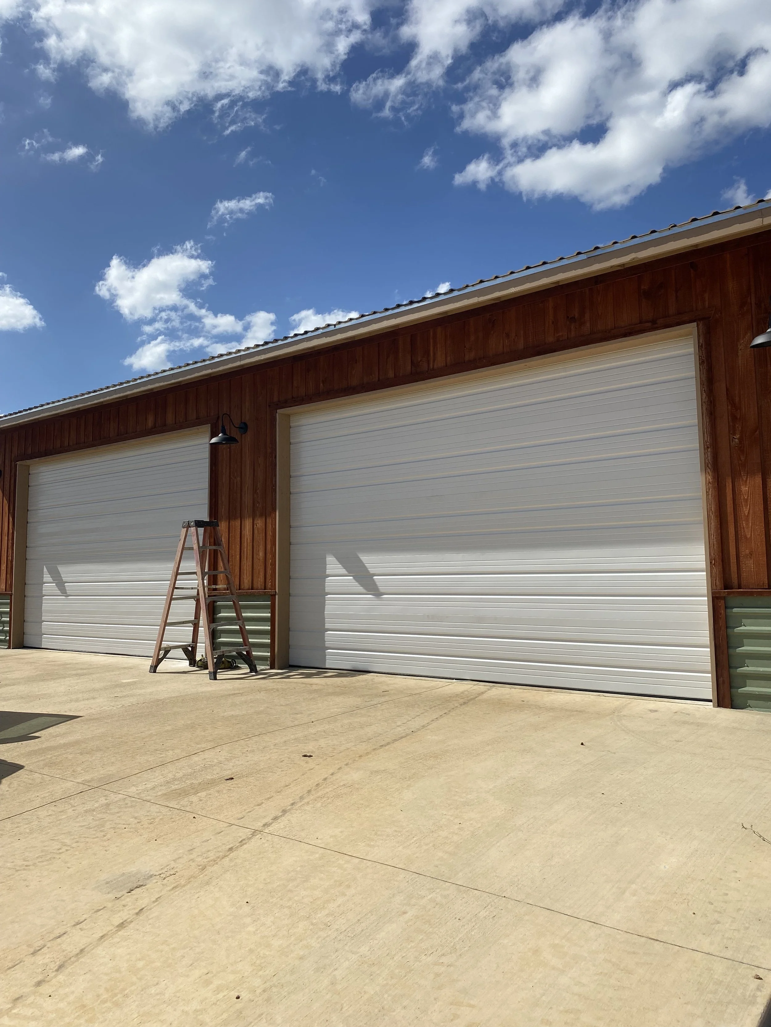 Two 16x12 white commercial garage doors on a barndominium building under a blue sky with scattered clouds; a ladder leaning against one of the building.