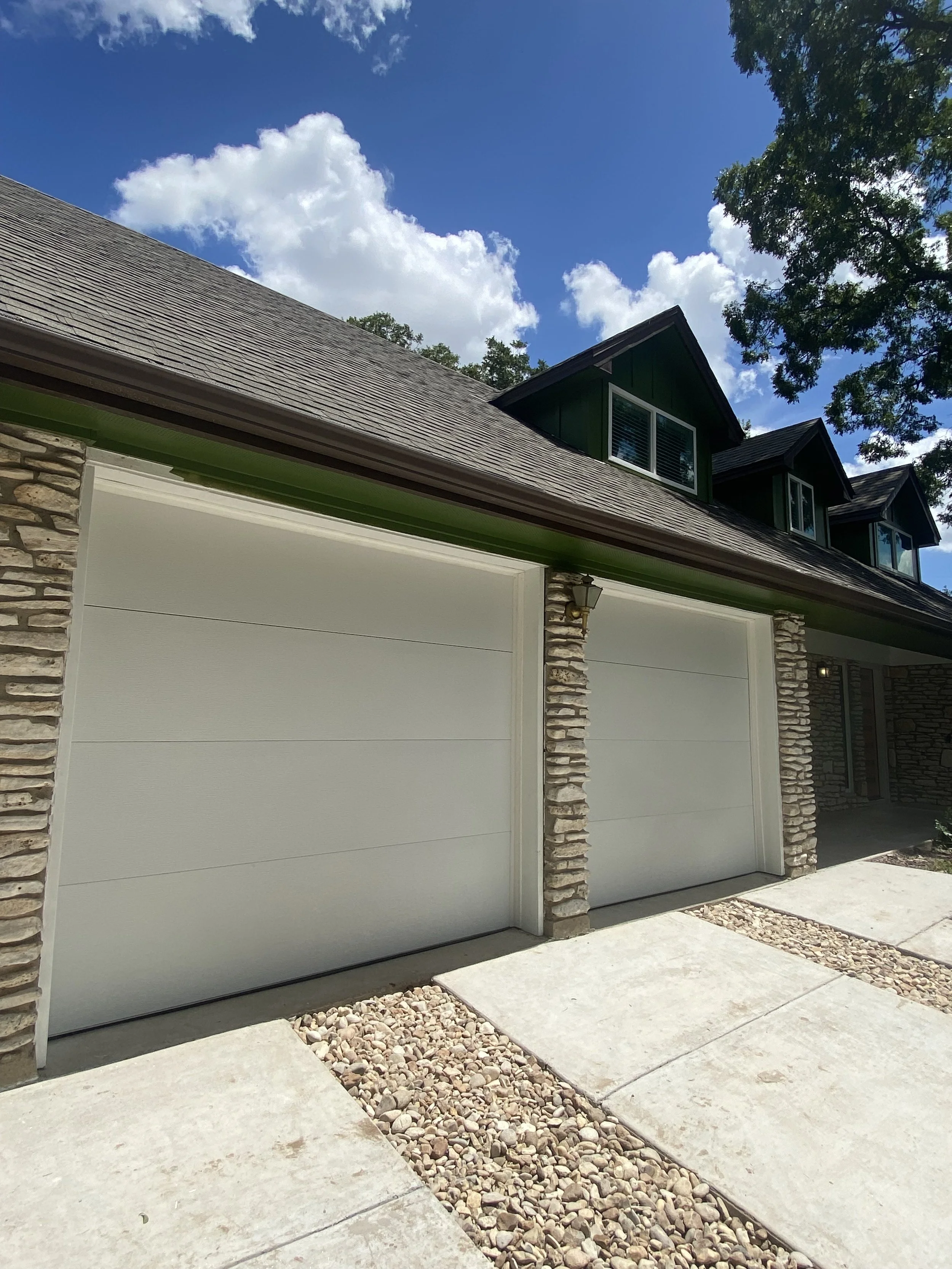 View of a modern house with two closed white flush garage doors, stone columns, a roof with shingles, and a sidewalk with decorative rocks in front. There are trees and a partly cloudy sky in the background.