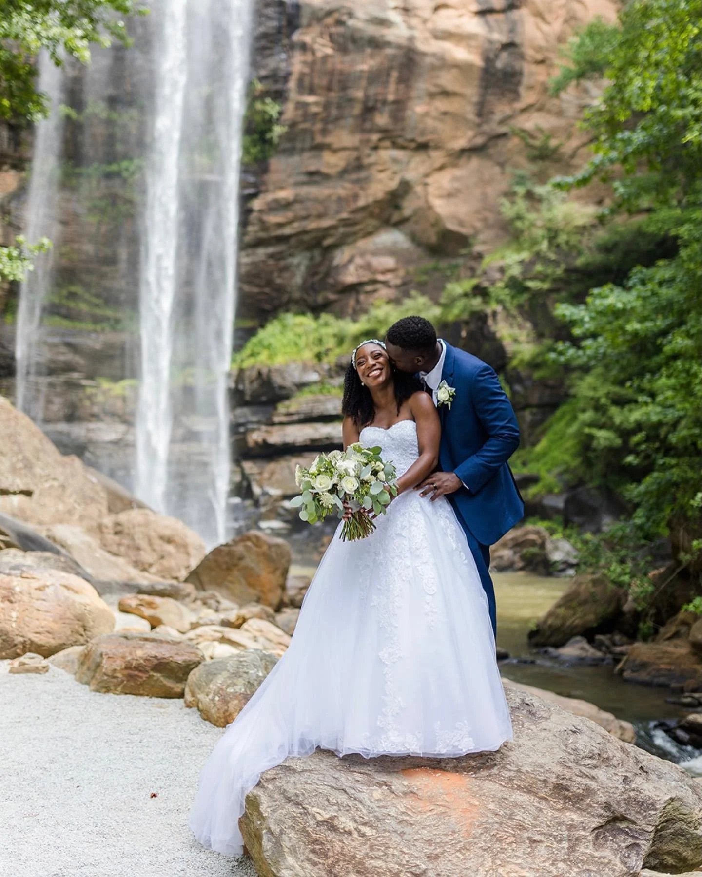 A newlywed couple in wedding attire standing on rocks near a waterfall, with greenery and a rocky cliff in the background.