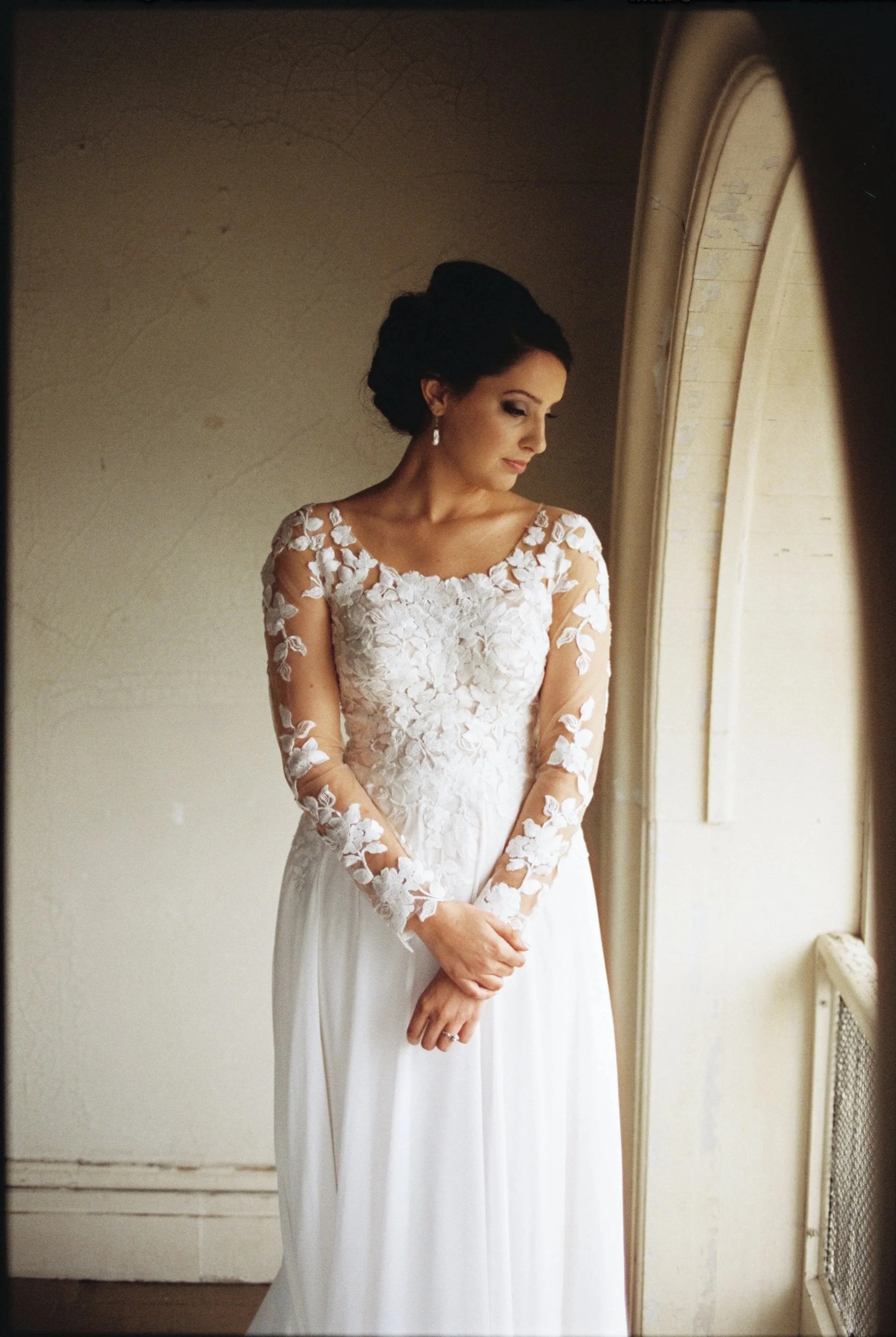 A bride in a white wedding dress with lace floral embroidery on the sleeves and bodice, standing by a window and looking down.
