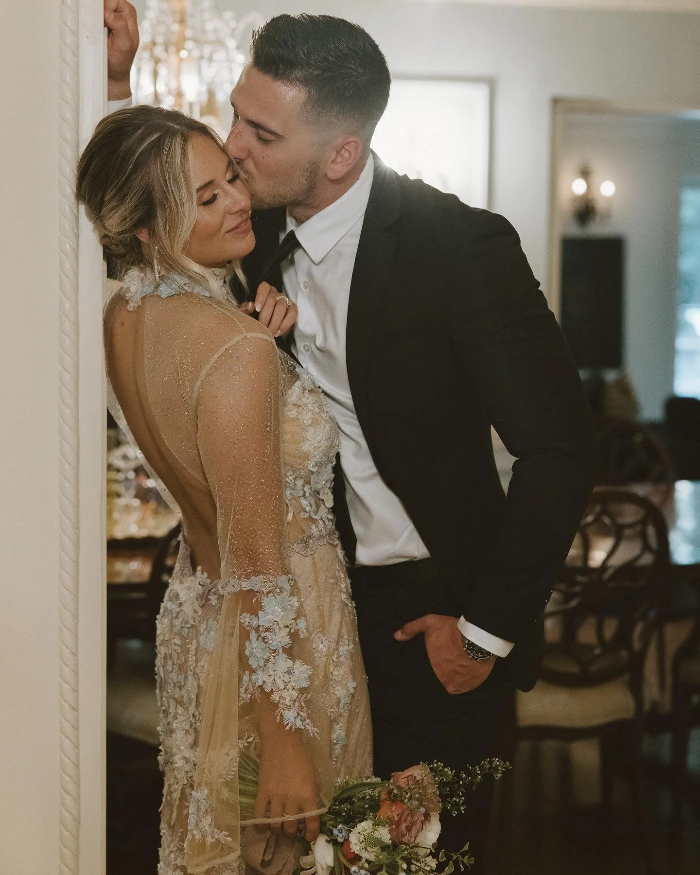 A man and woman are sharing an intimate moment in a warmly lit room. The woman is dressed in an elegant, sheer, lace dress and holding a bouquet, while the man is in a black tuxedo with a white shirt. He is kissing her on the forehead as she leans into him with her eyes closed.