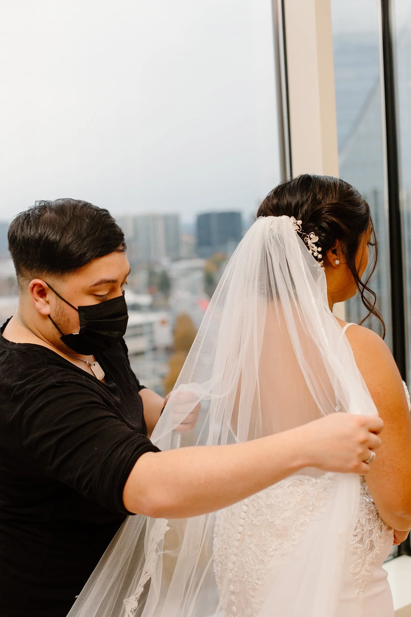 A bride is getting ready with the help of a stylist or assistant, who is attaching her veil. The bride is wearing a white wedding dress with lace details, and has dark hair styled with a decorative hairpiece. The assistant is wearing a black face mask and black clothing, and is adjusting the veil on the bride's head near a tall window with a cityscape view outside.