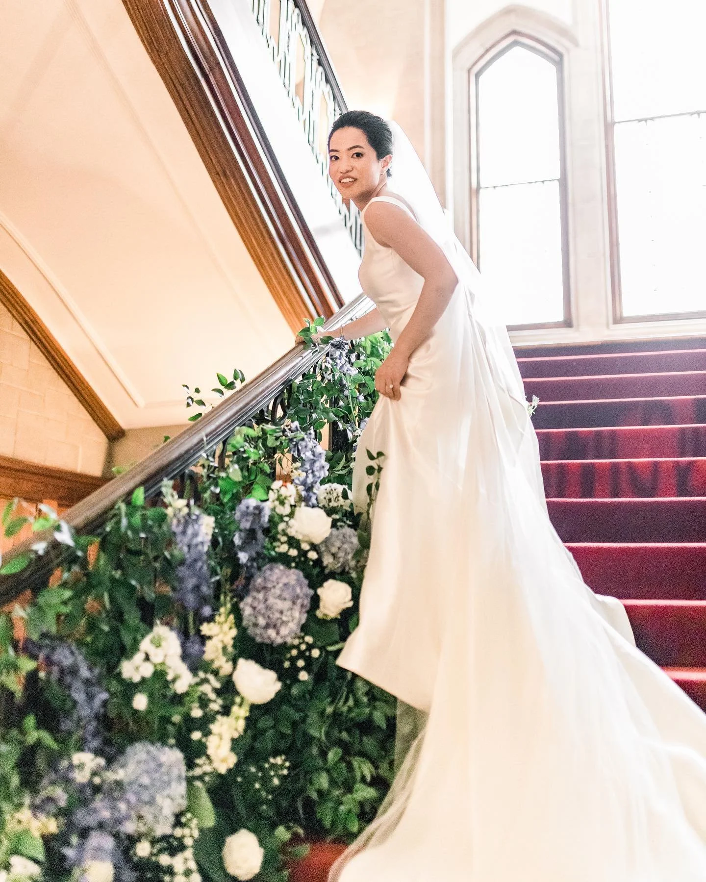 A bride in a white wedding gown standing on a red-carpeted staircase, decorated with flowers and greenery, holding the skirt of her gown, inside a building with high windows.