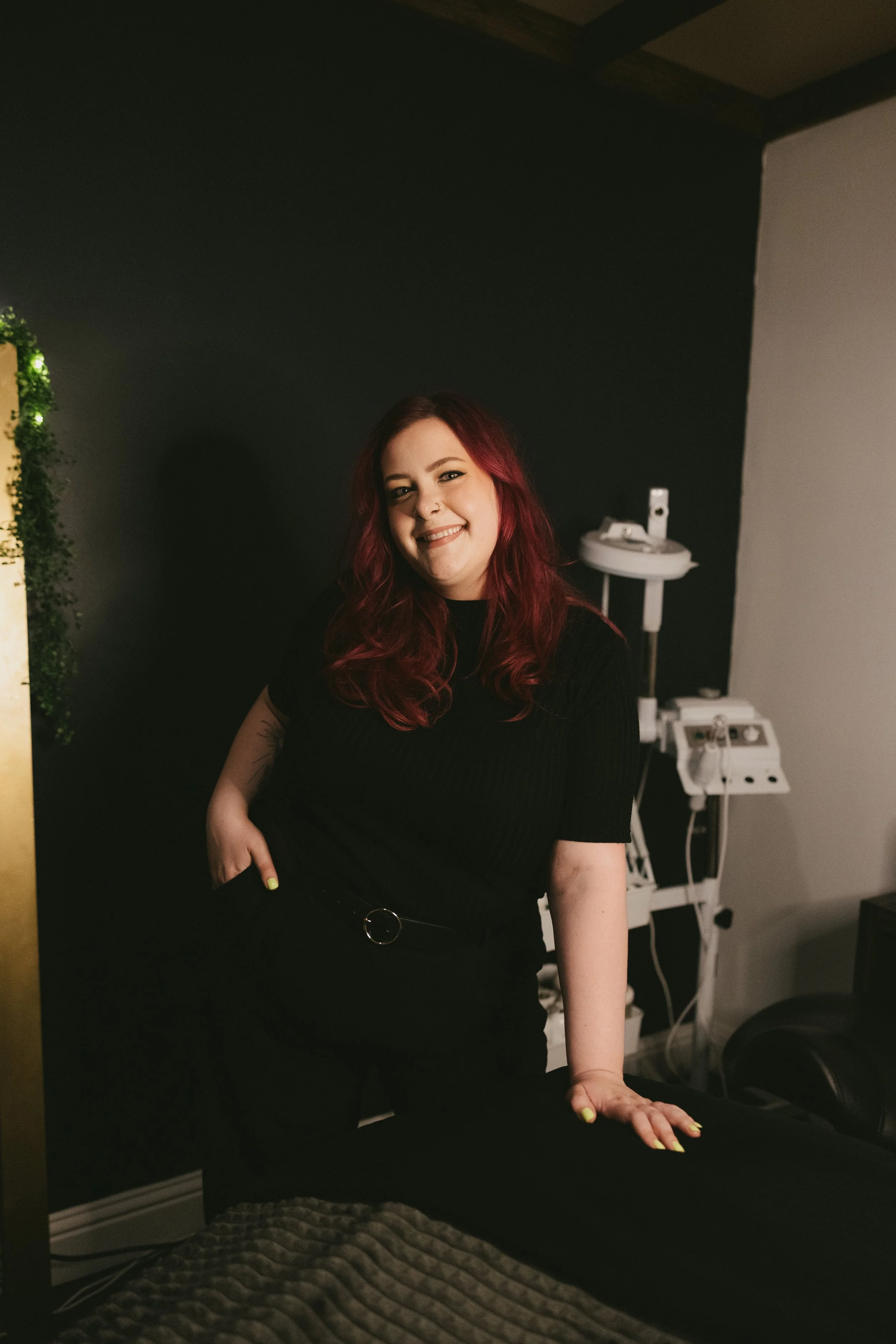 A woman with long red hair wearing a black shirt and black pants, leaning with her hand on a black surface, smiling at the camera in a dimly lit room.