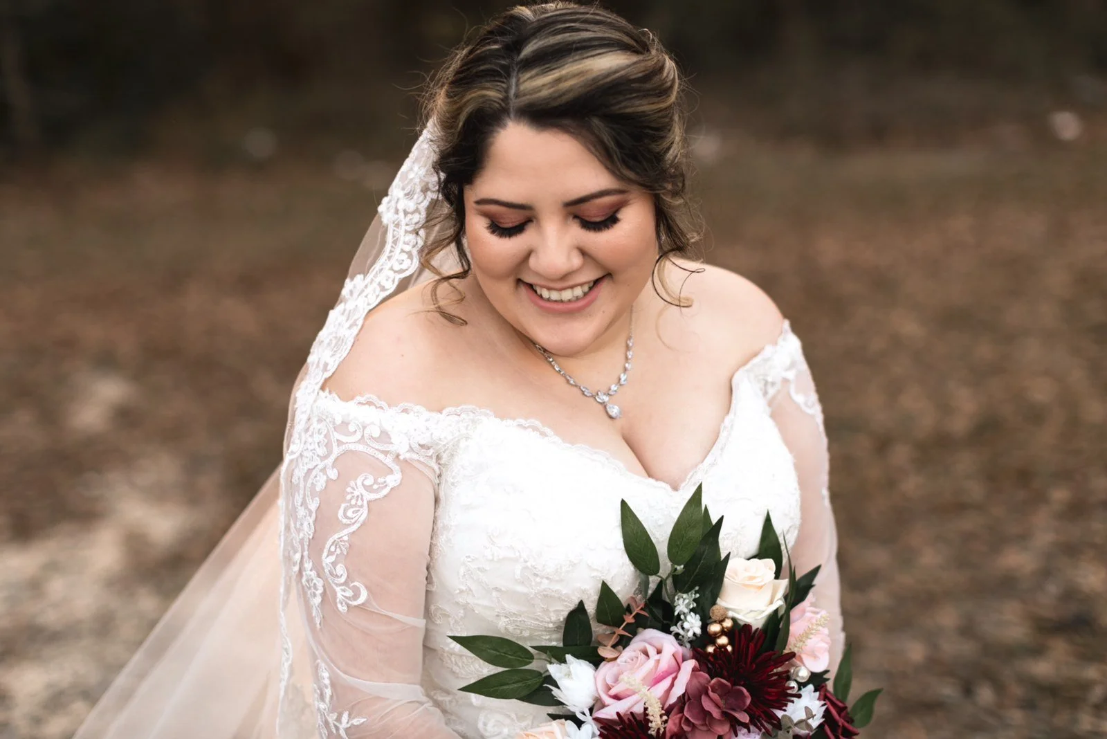 A smiling bride in a white lace wedding dress holding a bouquet of pink, burgundy, and white flowers.