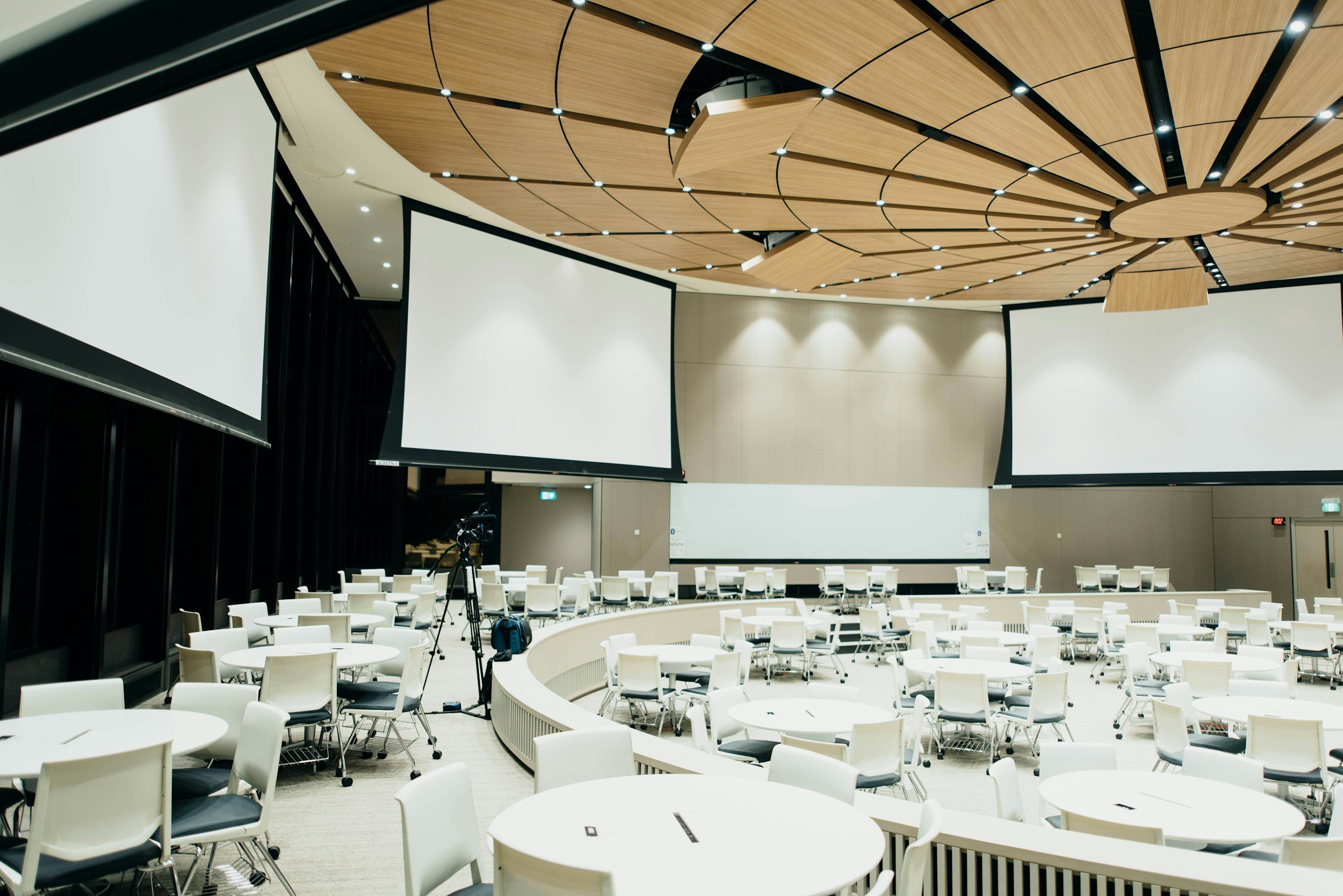 A modern conference room with multiple large projection screens, white chairs, and round tables arranged in a curved pattern, illuminated by ceiling lights.