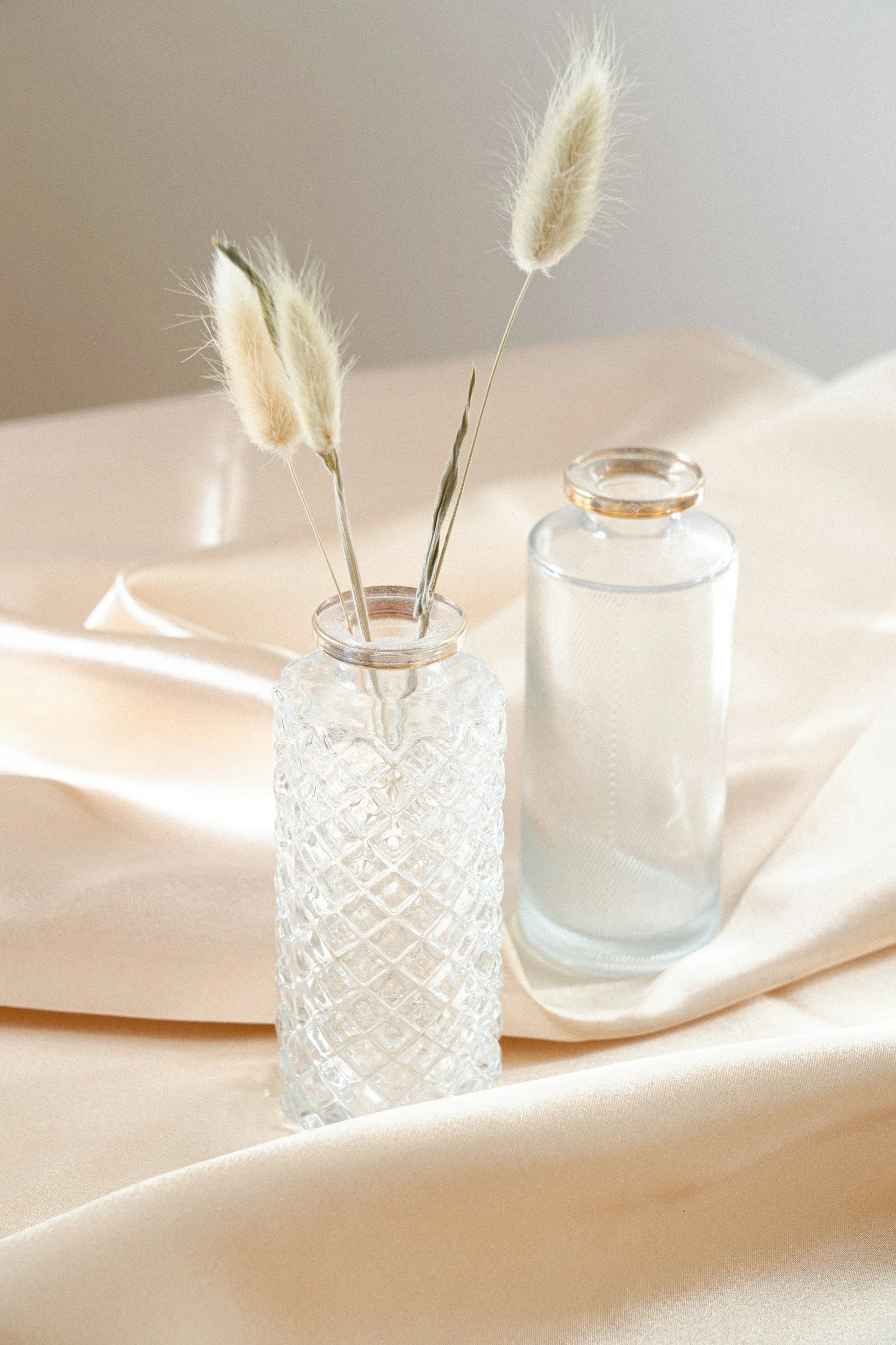 Two glass vases, one with a diamond pattern and the other clear, are placed on a soft beige fabric. The patterned vase contains dried pampas grass. The scene is softly lit with a neutral background.