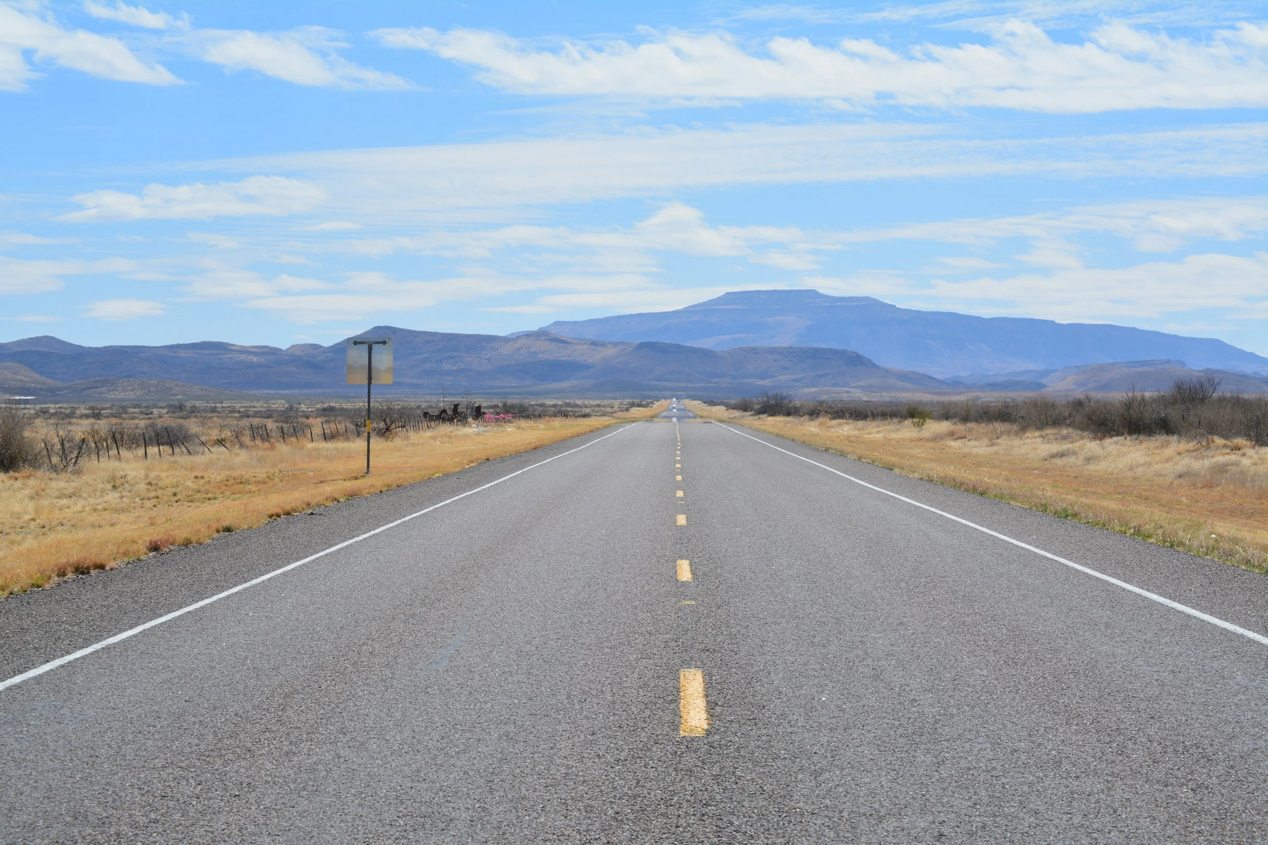 A straight road extending into the distance through a desert landscape with mountains in the background, under a blue sky with scattered clouds.