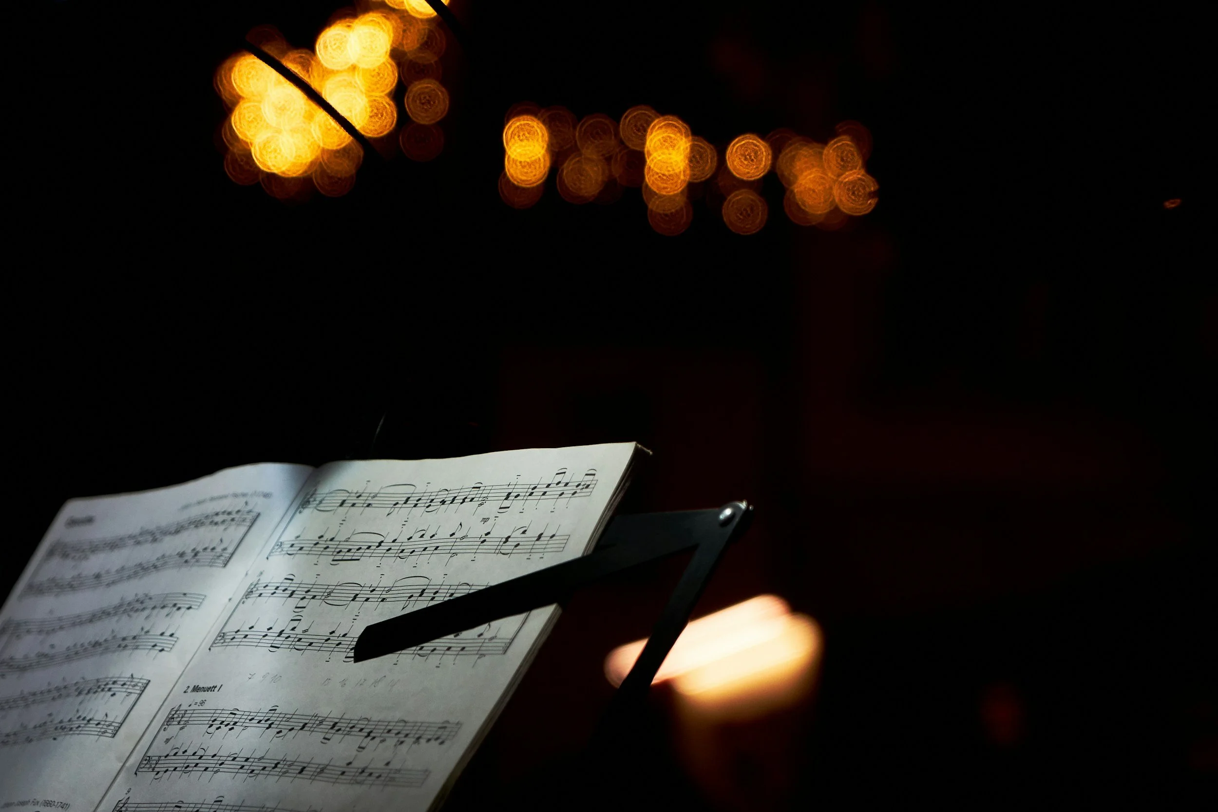 Sheet music on a stand illuminated by warm stage lighting, with blurred yellow lights in the background.