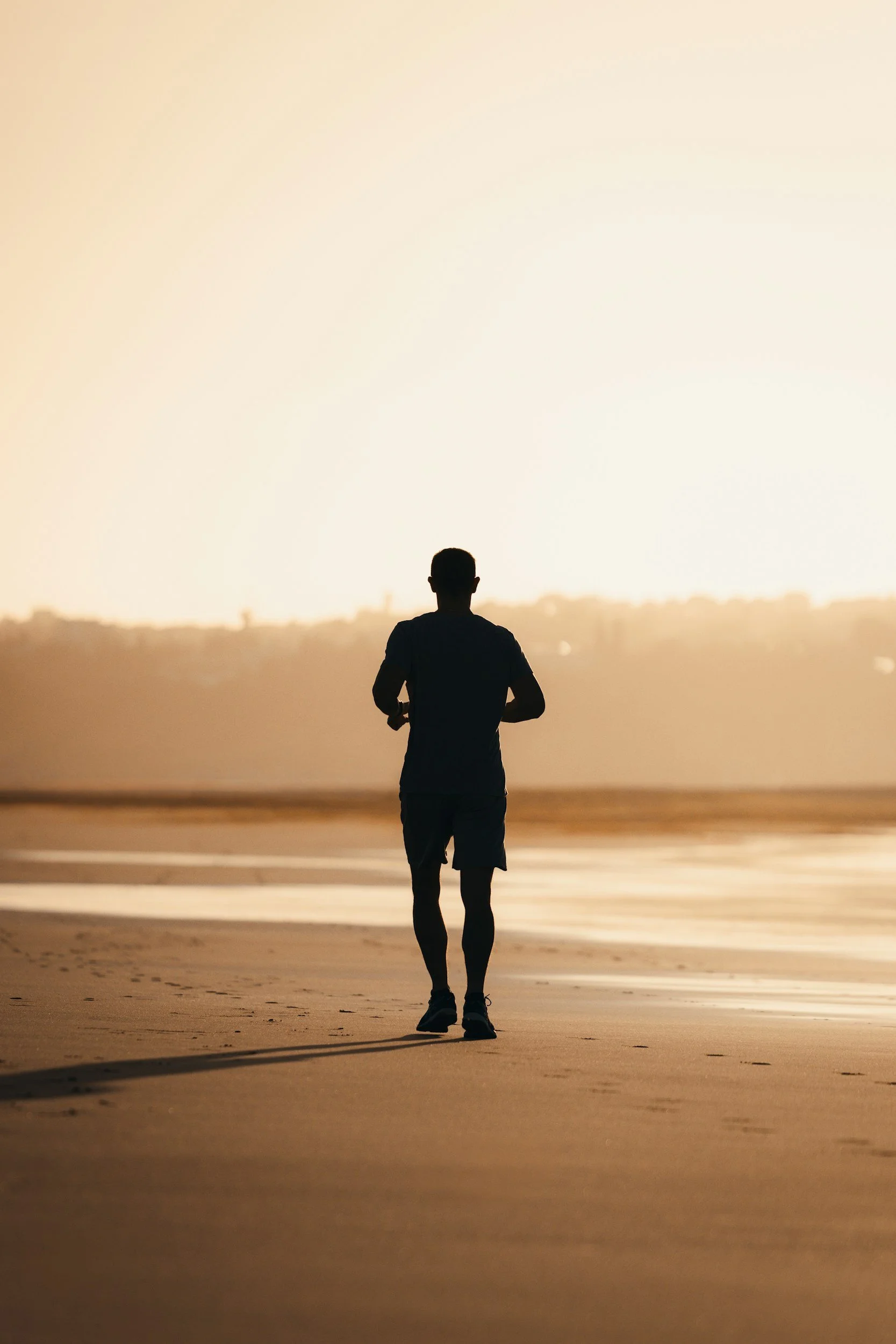 A person jogging on a sandy beach at sunset or sunrise with a light yellow sky and distant cityscape.