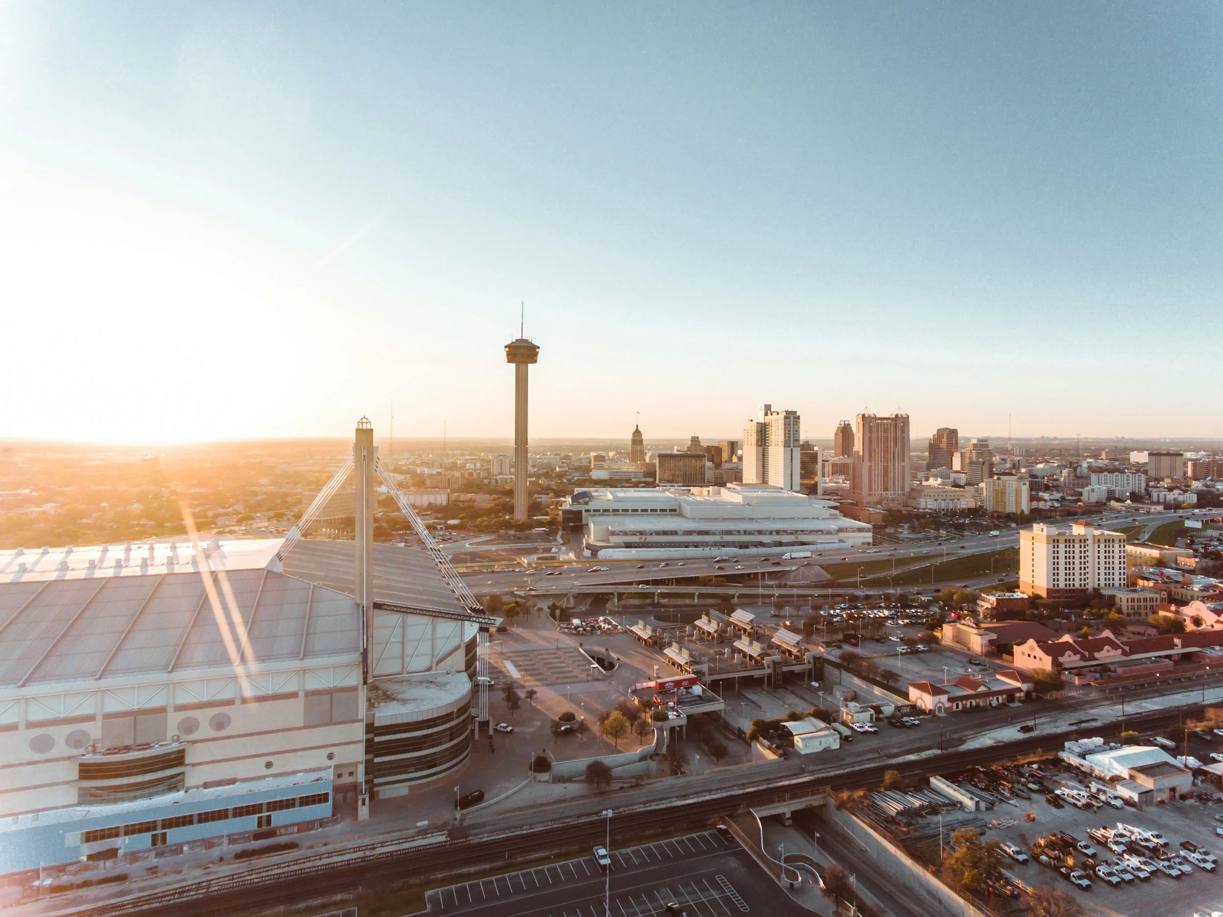 A city skyline at sunset with tall buildings, including a tower, and a large sports arena in the foreground. Streets and parking lots are visible below.