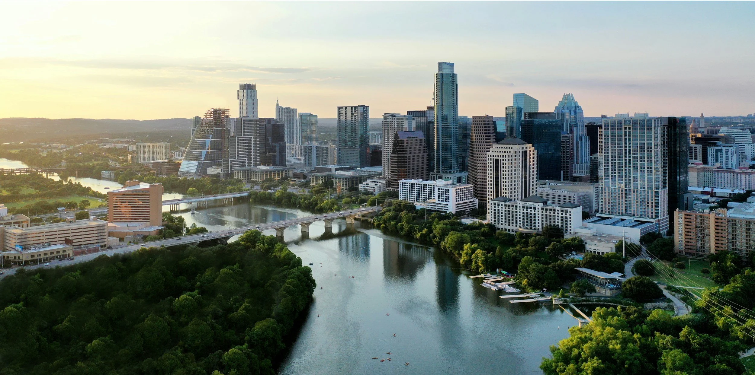 Aerial view of downtown Austin, Texas, skyline with tall buildings and the Colorado River in the foreground during sunset.