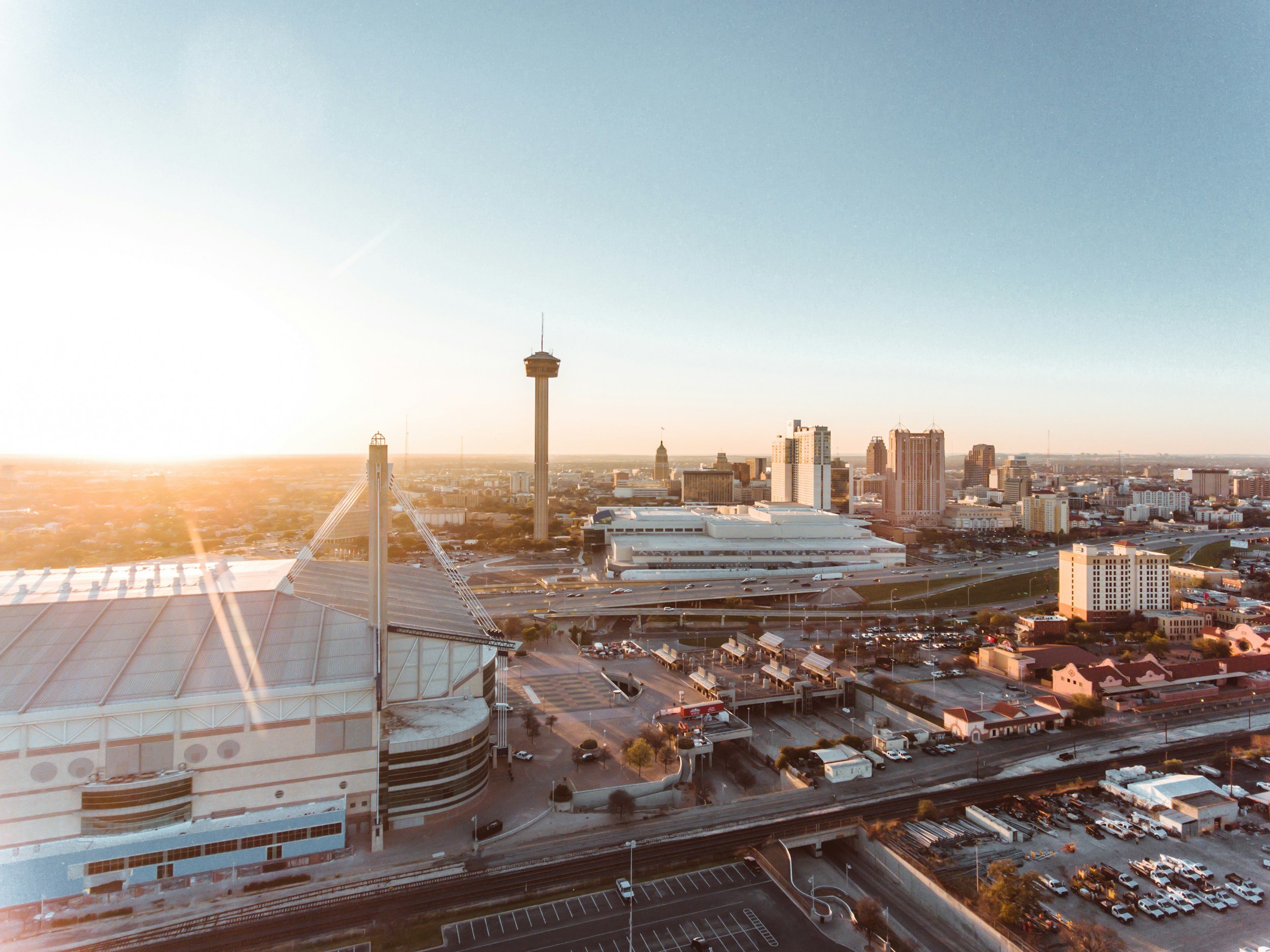 San Antonio skyline at sunset with several high-rise buildings, including a prominent tower with an observation deck, and a stadium with a curved roof in the foreground. The sky is clear with a gradient from light to dark blue.