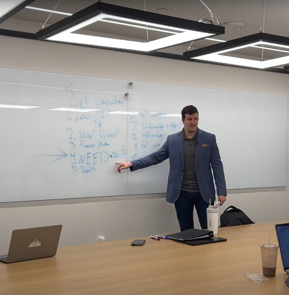 Alex Zabaski in a blue blazer standing in front of a whiteboard, teaching sales, in a conference room with a live audience.