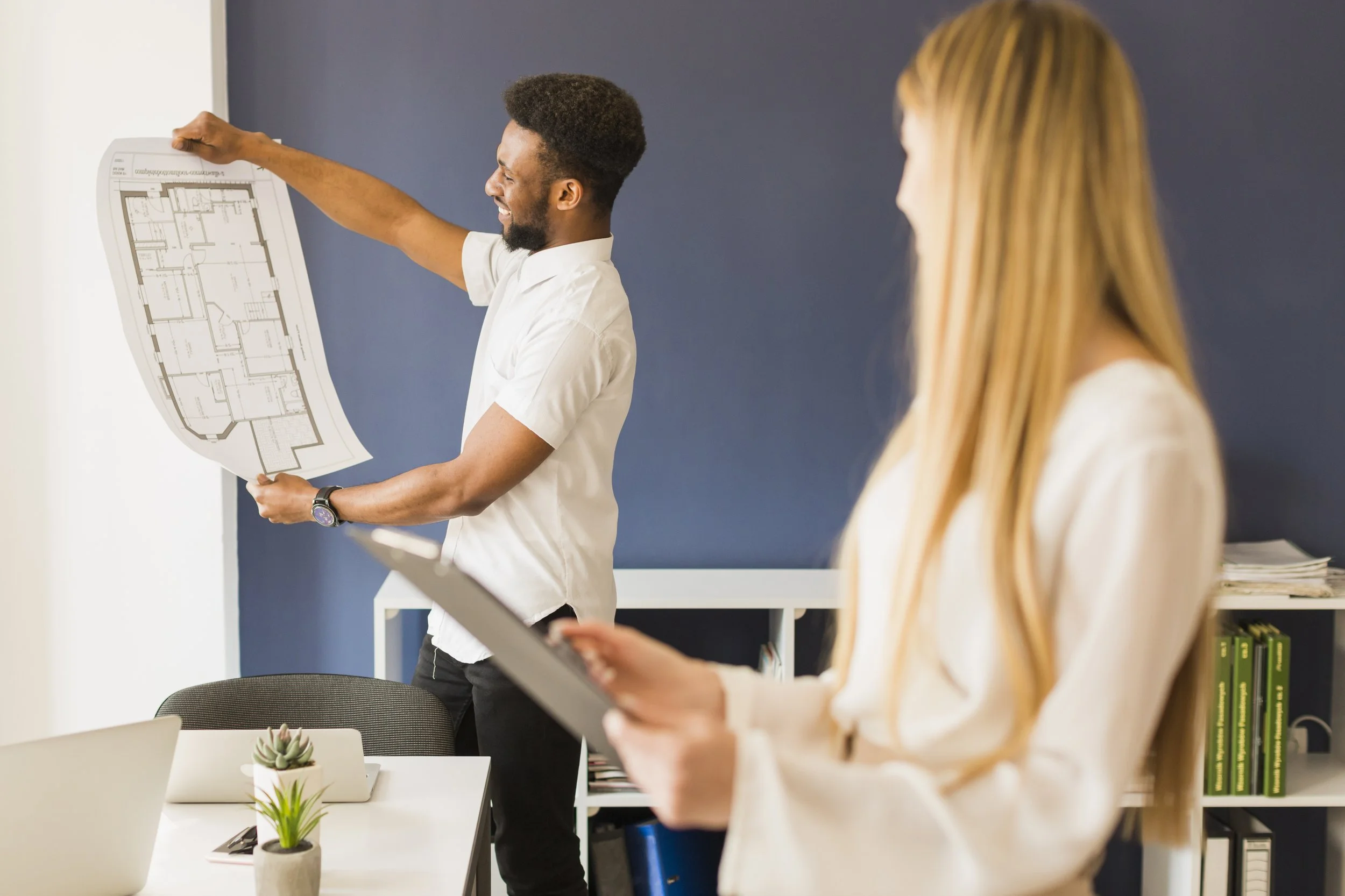A man presenting a remodeling blueprints to a woman holding a tablet in a modern home with a dark blue accent wall and a small plant on the table.