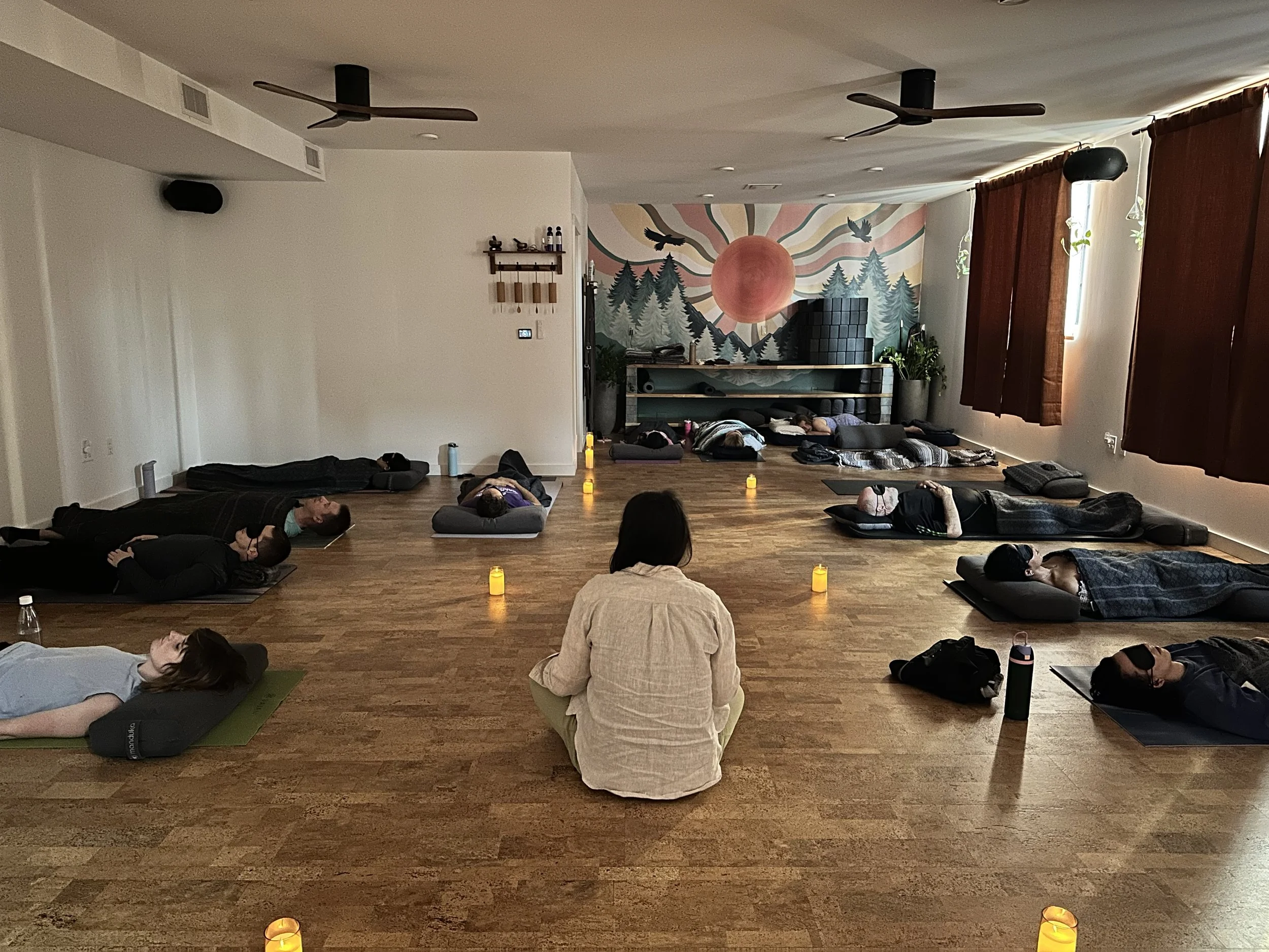 People lying on yoga mats in a meditation room, with candles on the floor, a mural of a sunset over mountains, and a woman sitting cross-legged in the foreground.