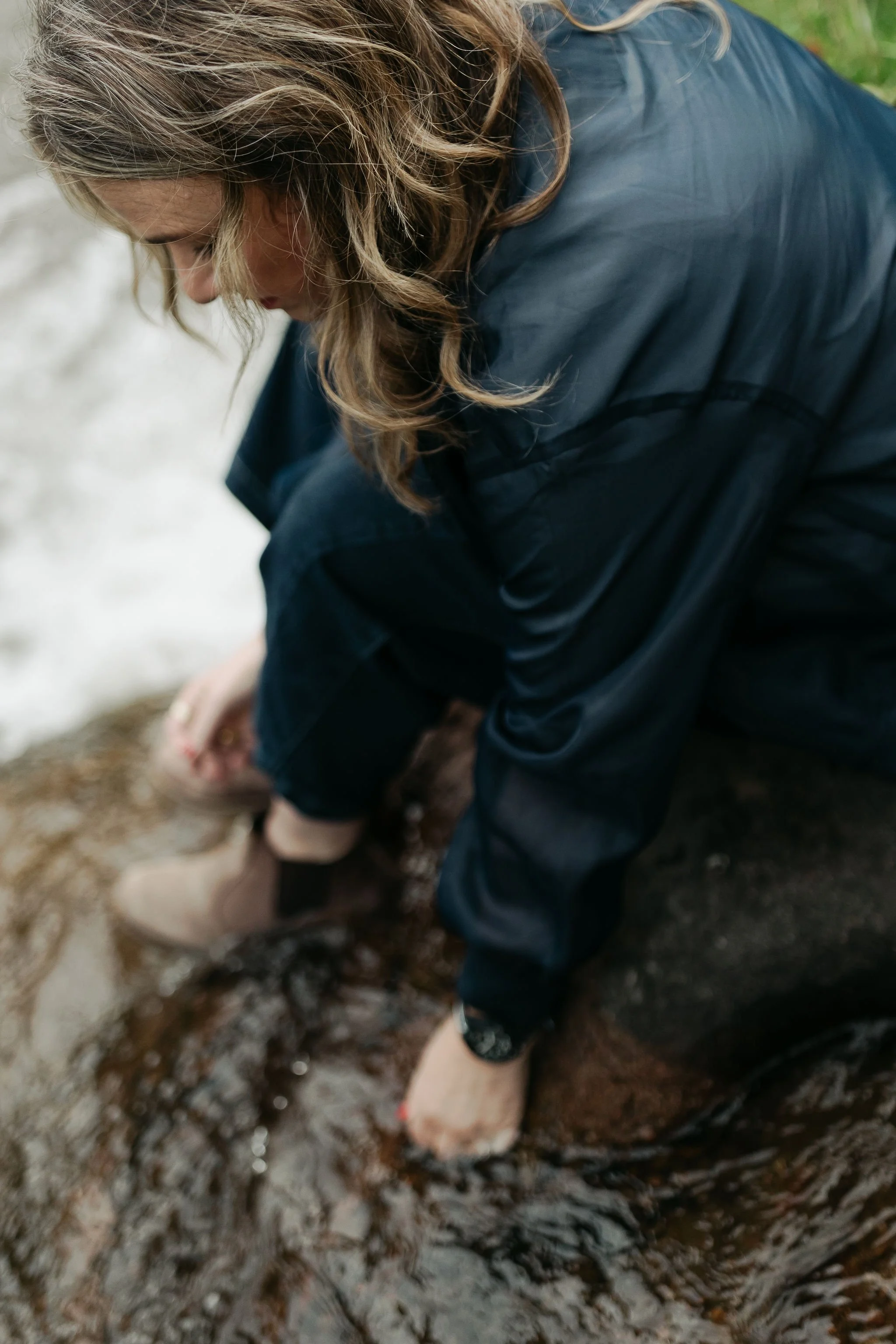 A person wearing a dark rain jacket is outdoors, crouching on wet rocks near water, with their hands submerged in water.