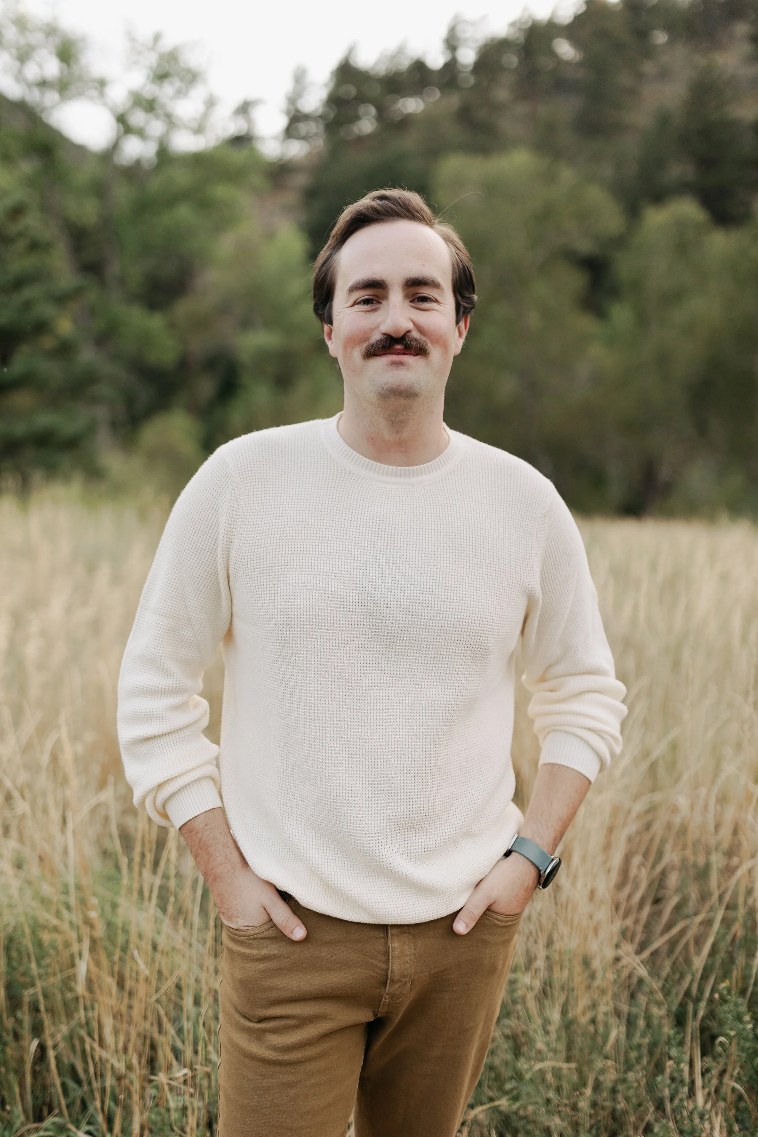 Gabriel Krush standing outdoors in a field of tall, yellow grass with trees in the background, wearing a cream-colored sweater, brown pants, and a watch.