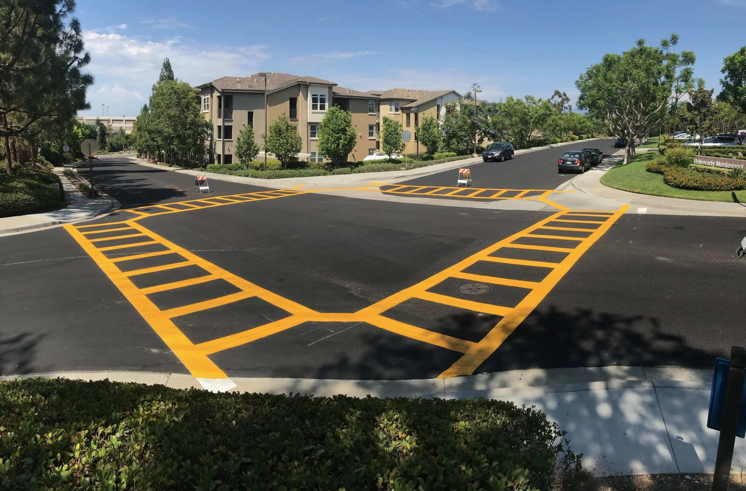 A traffic cross walk with freshly painted yellow striped no-parking zones and barriers blocking part of the area, surrounded by trees, grass, and residential buildings.