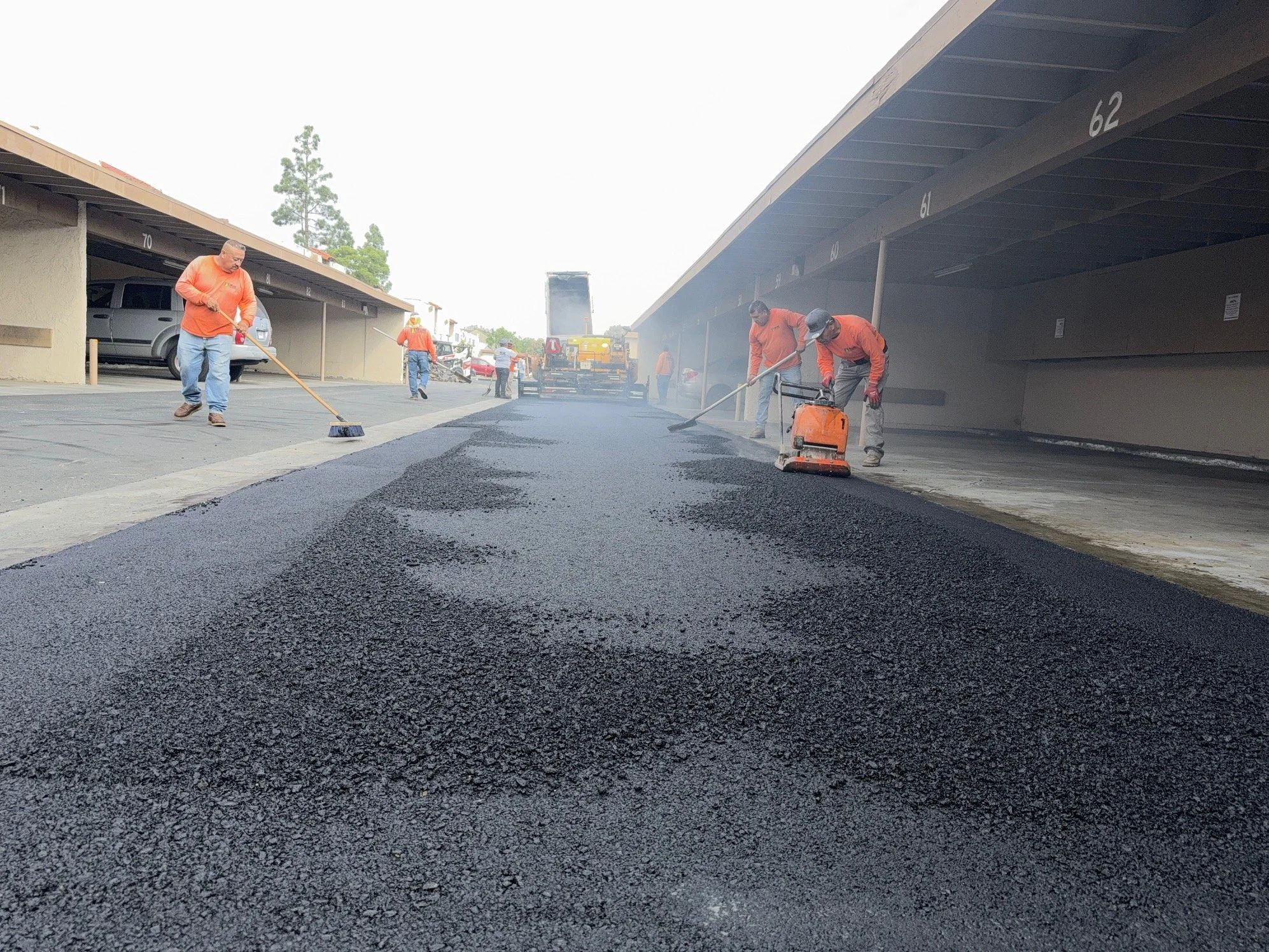 Workers are paving a parking lot with asphalt under a carport. They are using shovels, tools, and machinery to spread and smooth the asphalt.