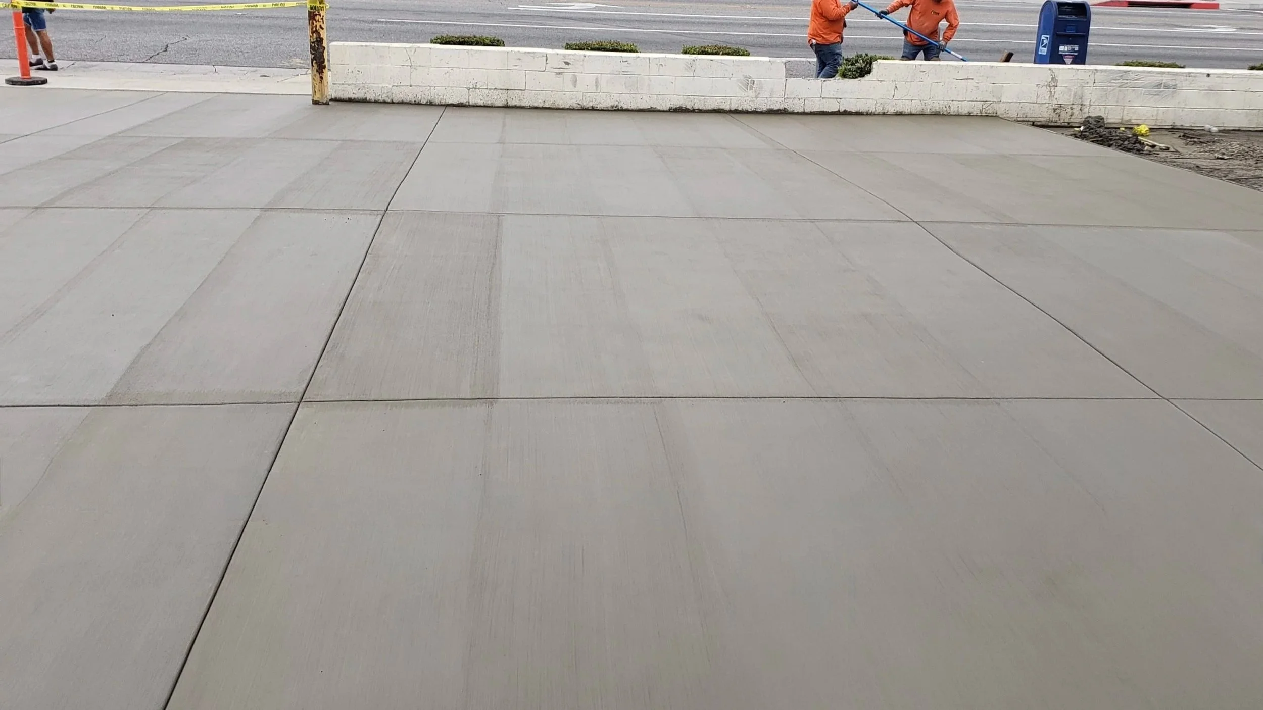 Newly laid concrete sidewalk with construction workers working on the adjacent road in the background.