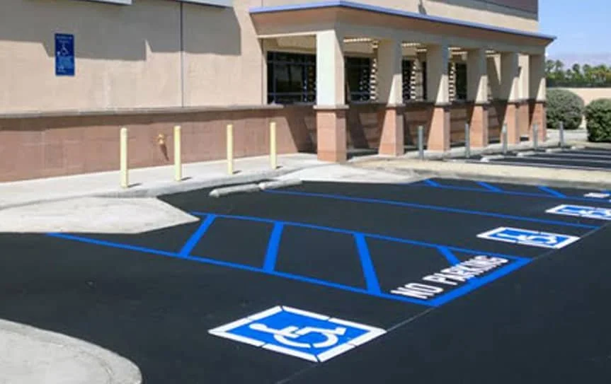 Empty handicapped parking spaces marked with blue lines and symbols, located outside a building with a pinkish facade and yellow vertical poles on sidewalk.