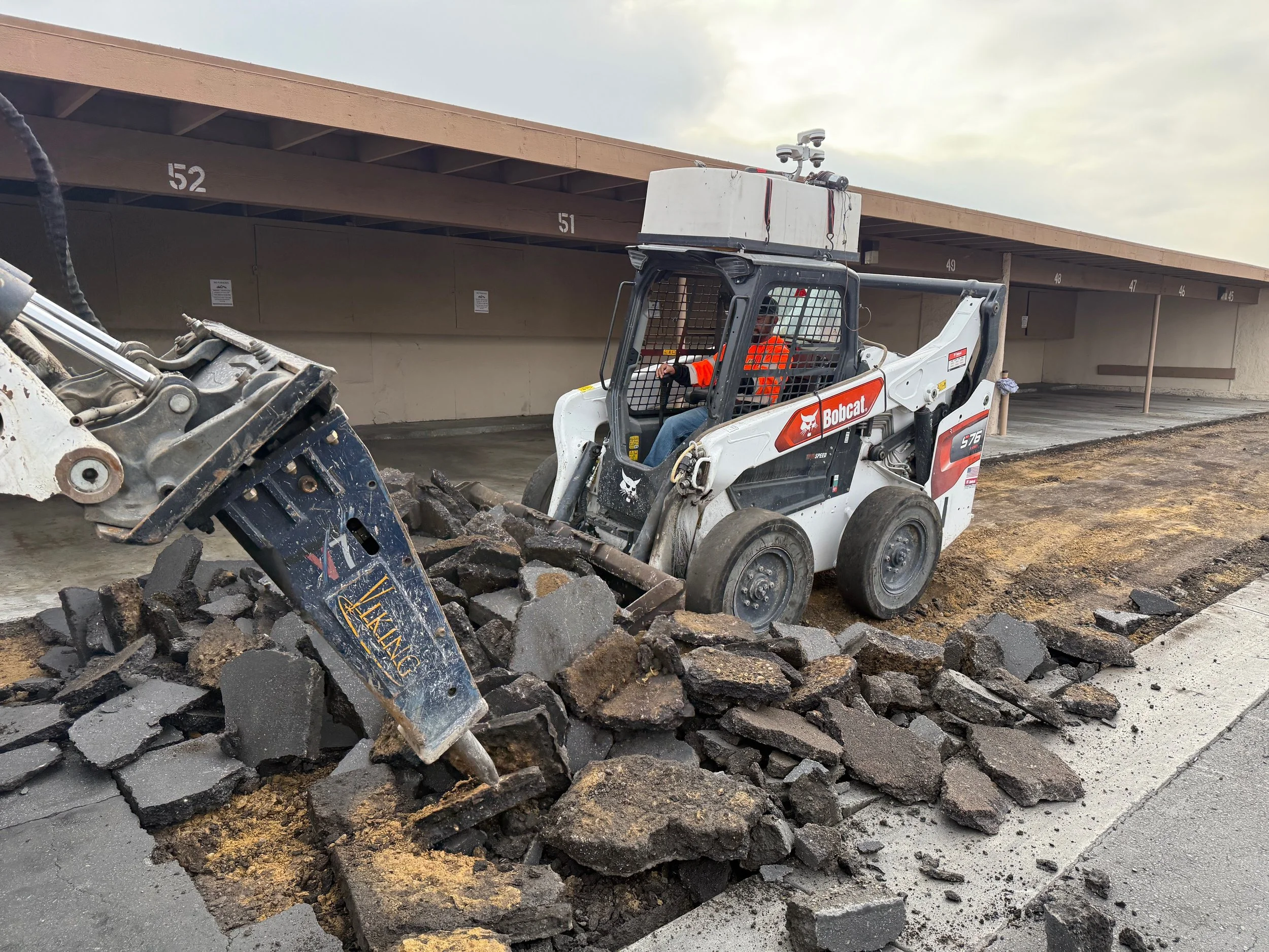 A Bobcat mini skid steer loader is breaking up pavement with a hydraulic hammer attachment in front of a parking garage.