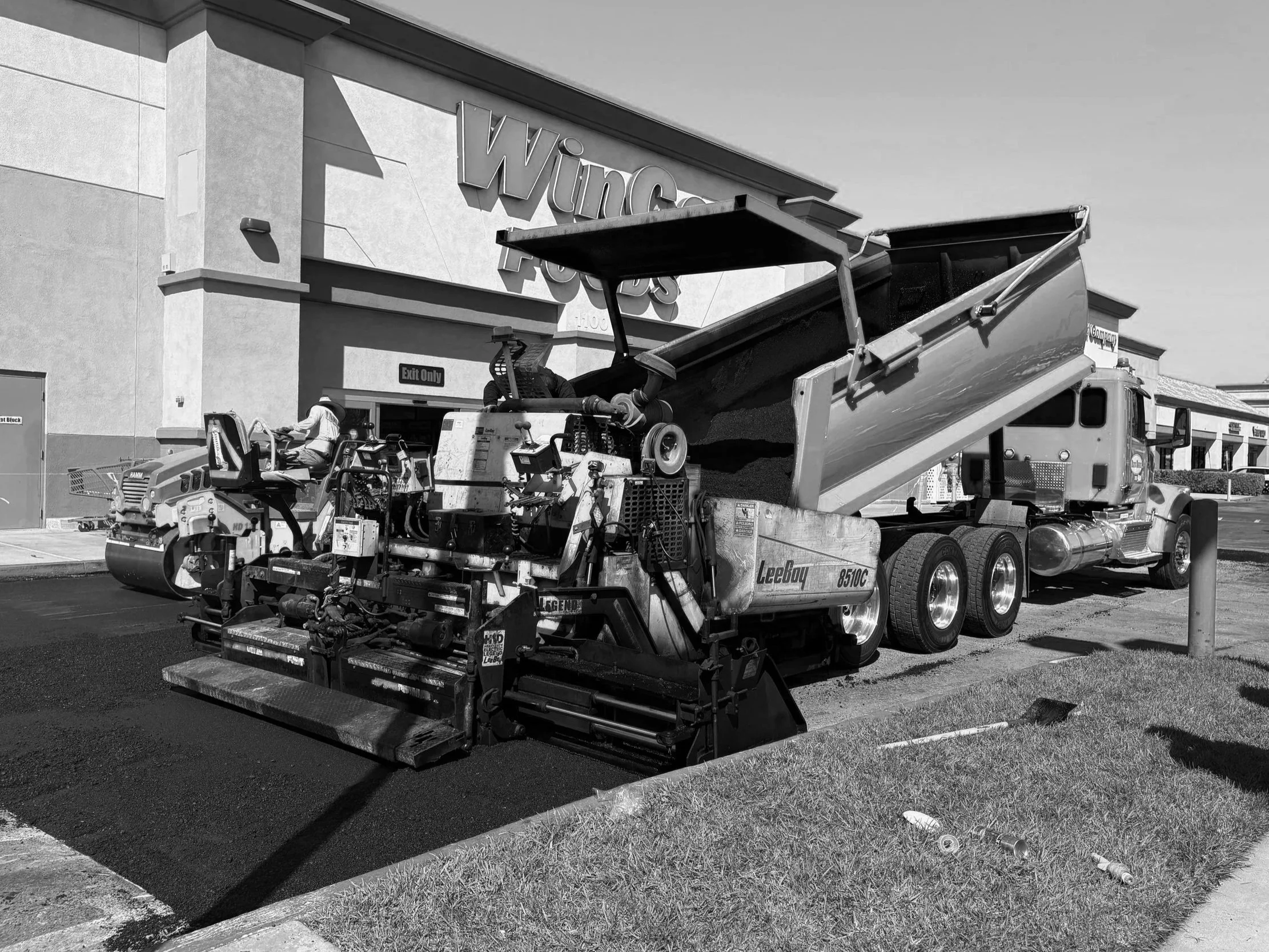 Construction equipment paving a parking lot. A large road paving machine is visible with asphalt being laid down, and a dump truck is working in tandem to unload asphalt into the paving machine.