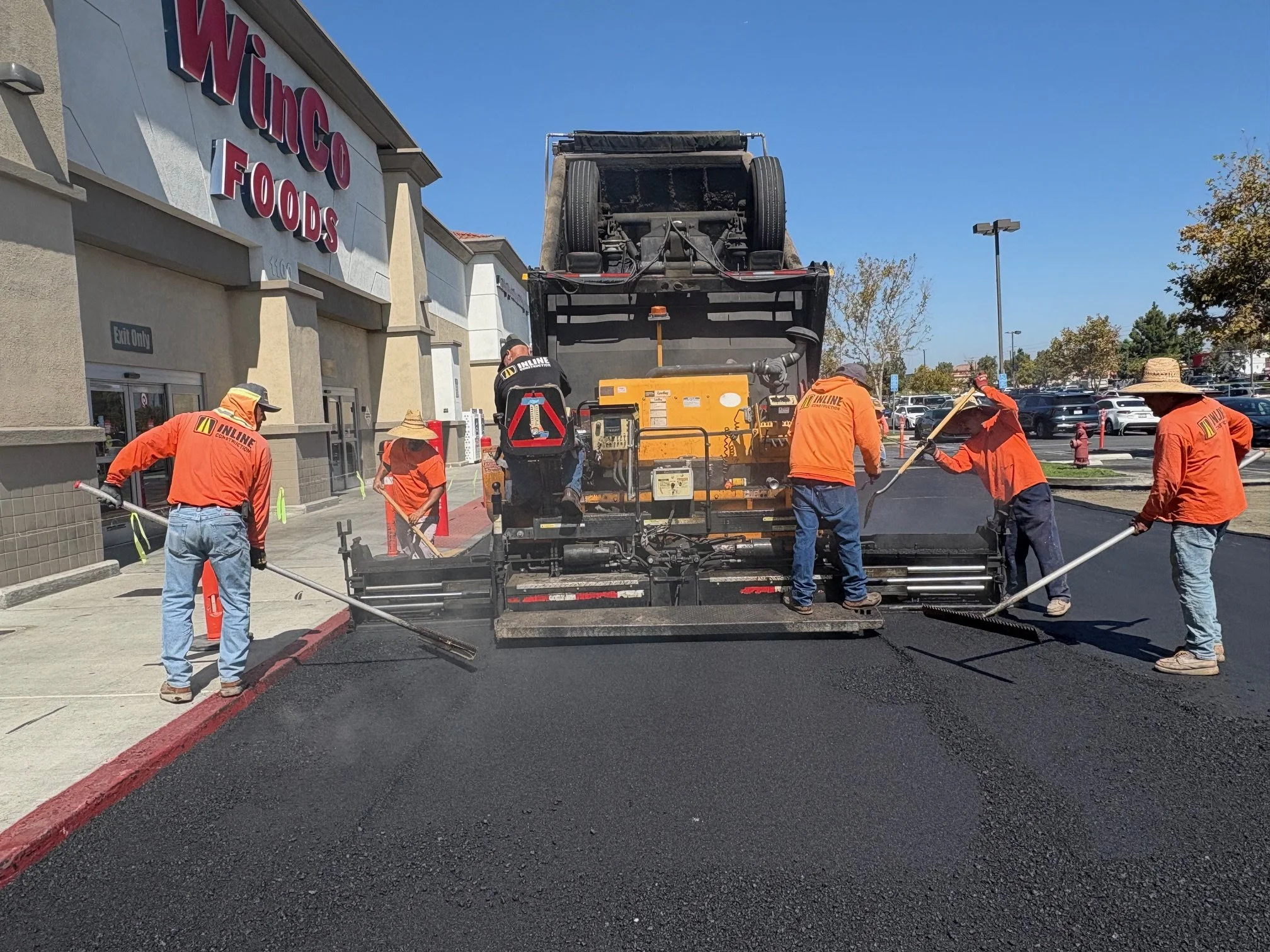 Workers wearing orange shirts are paving asphalt in a parking lot outside a grocery store called WinCo Foods. The workers are using shovels and rakes to spread the hot asphalt, which is freshly laid down. In the background, there are parked cars and trees under a clear blue sky.
