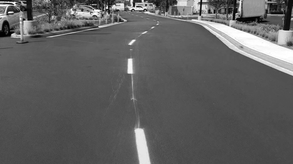 Empty city street with a cracked middle lane dividing line, parked cars on the sides, and buildings in the background.