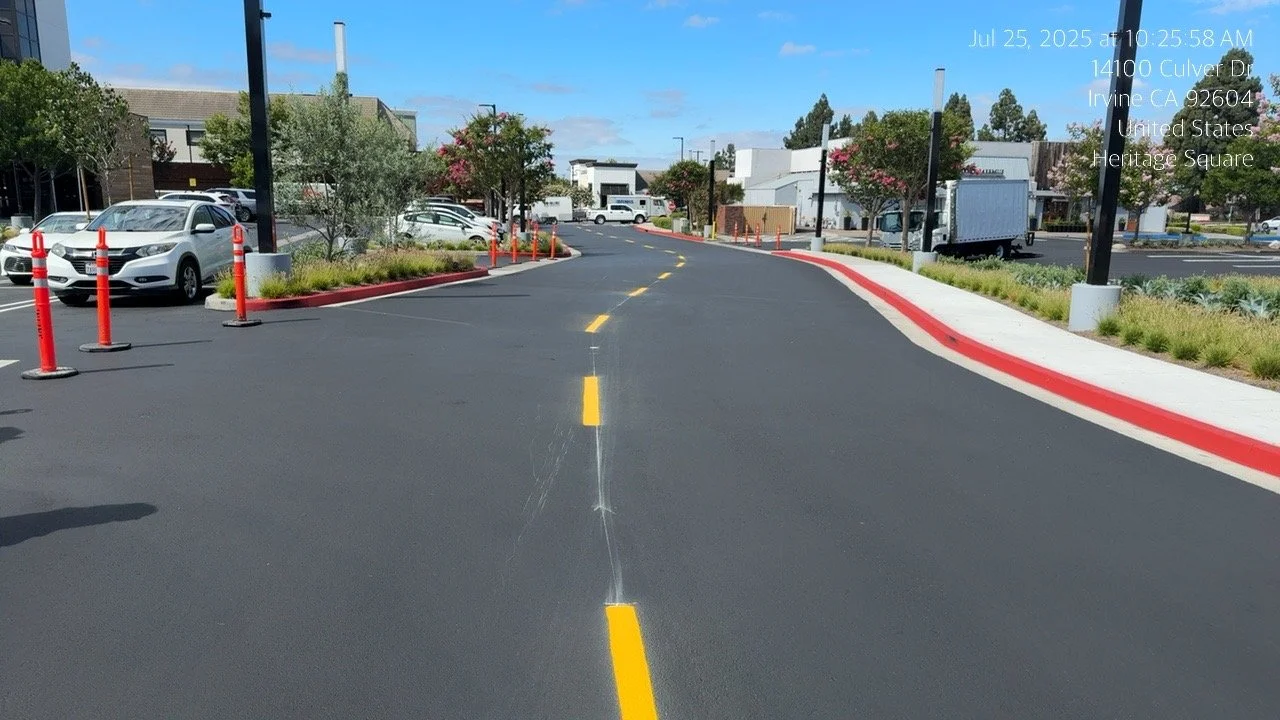 Empty parking lot with a cracked road, red curbs, and several parked cars. There are trees, buildings, and a truck in the background under a blue sky with some clouds. The photo has a timestamp and location overlay.