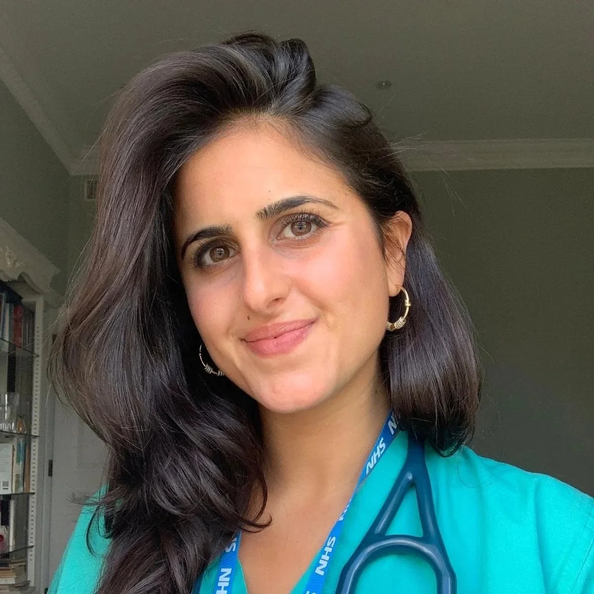 A woman with long dark hair, wearing a blue medical uniform, a blue lanyard, and a stethoscope around her neck, smiling indoors.