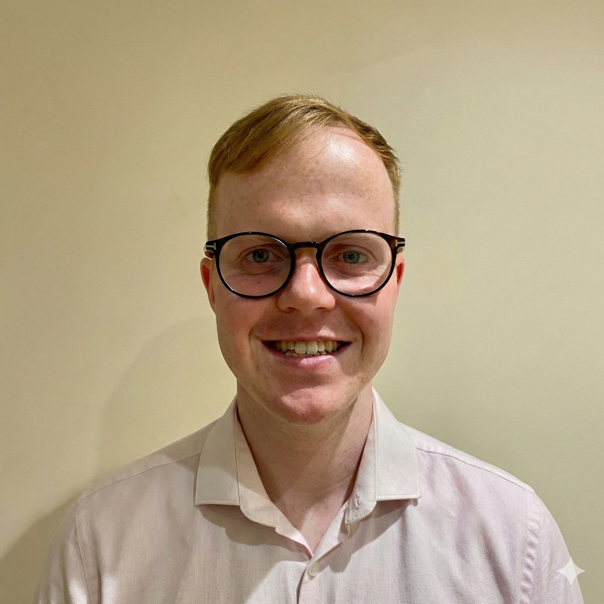 A smiling young man with red hair and glasses wearing a light-colored collared shirt against a plain beige wall.