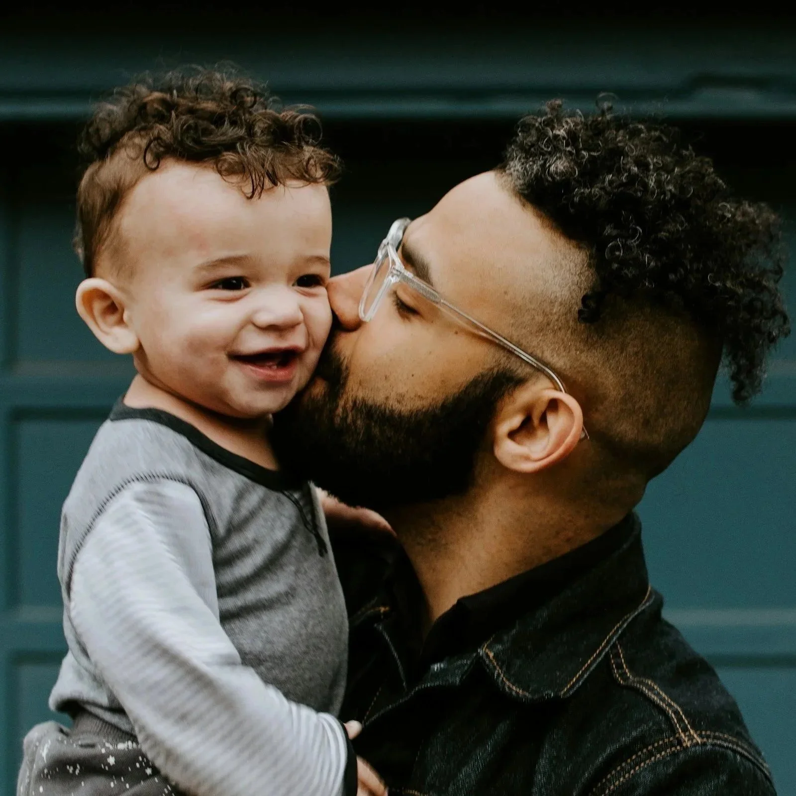 A man with glasses and a beard kissing a young boy on the cheek, both smiling. The boy has curly hair and is wearing a gray shirt with black trim, and is being held in the man's arms. They stand in front of a blue garage door.