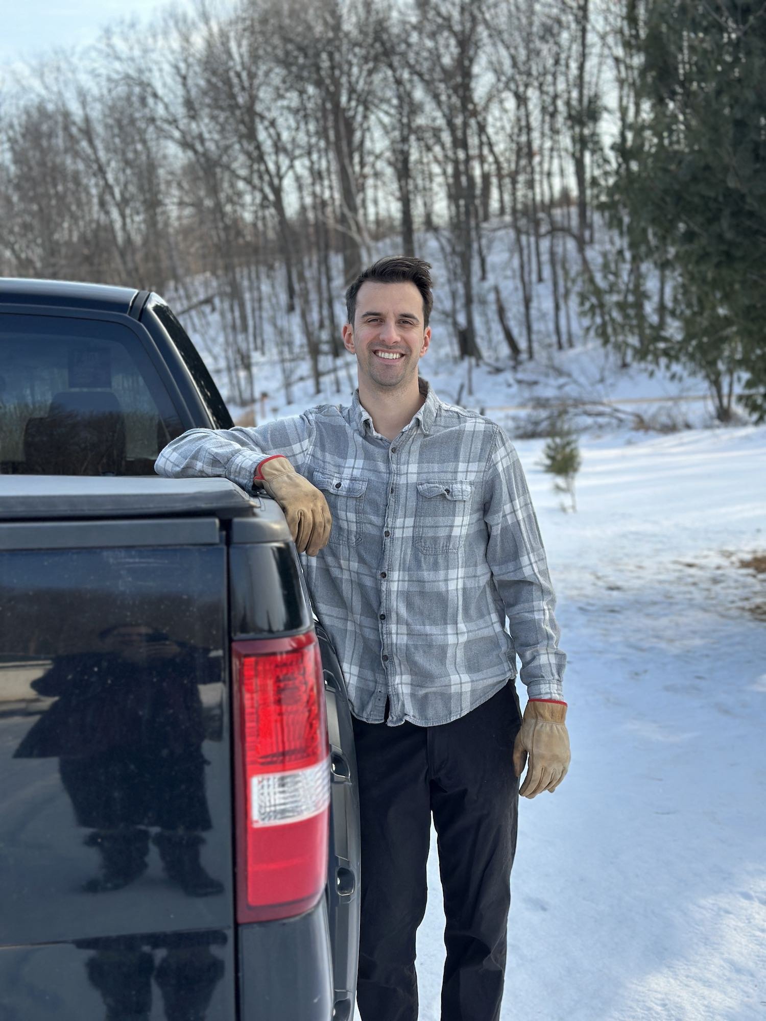 A man smiling standing in snow next to a black pickup truck, wearing a plaid shirt, black pants, and tan work gloves, with a wooded area in the background.